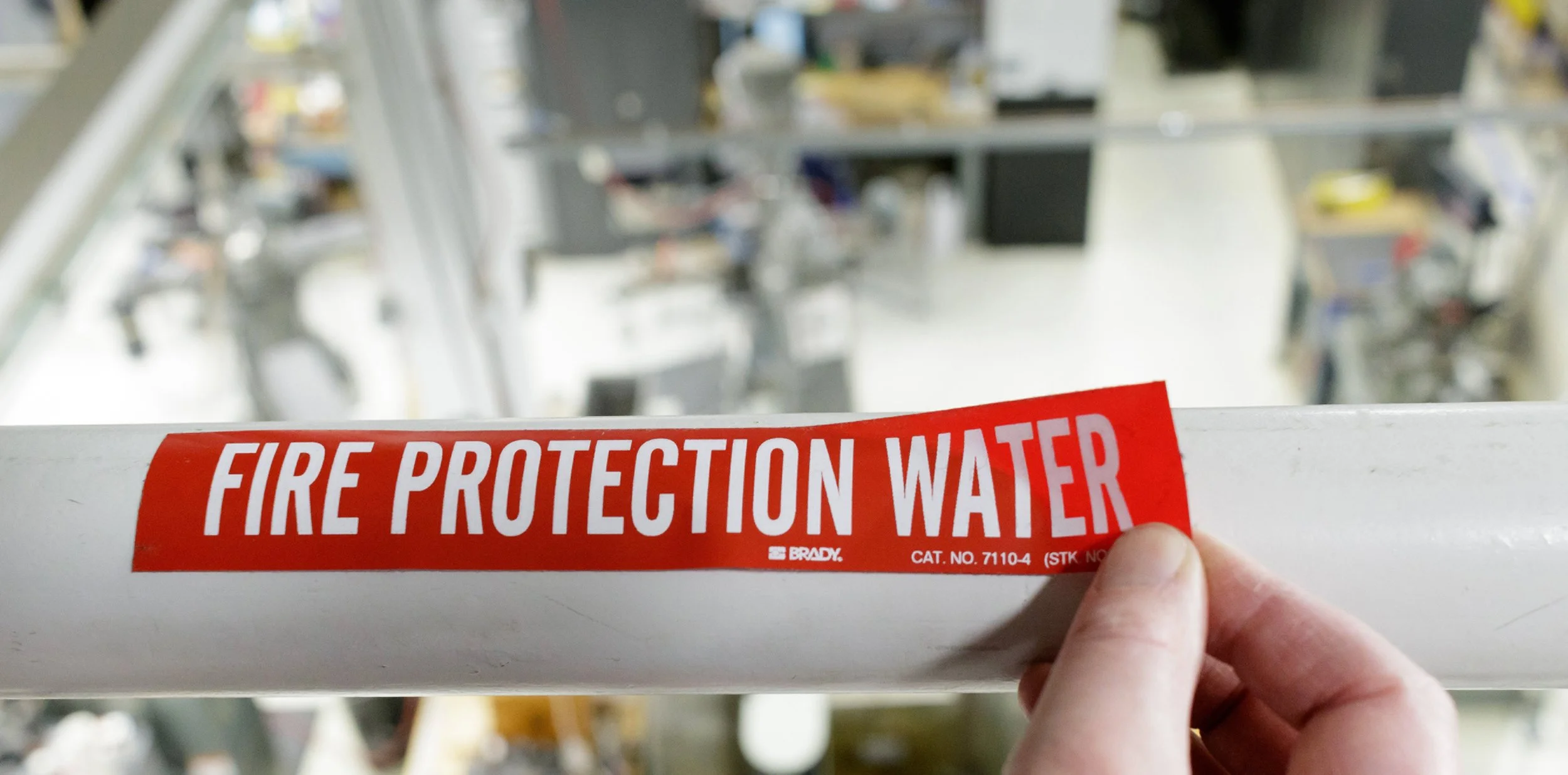 A hand holding a red and white label reading 'FIRE PROTECTION WATER' in front of a sprinkler system in a warehouse or industrial setting.