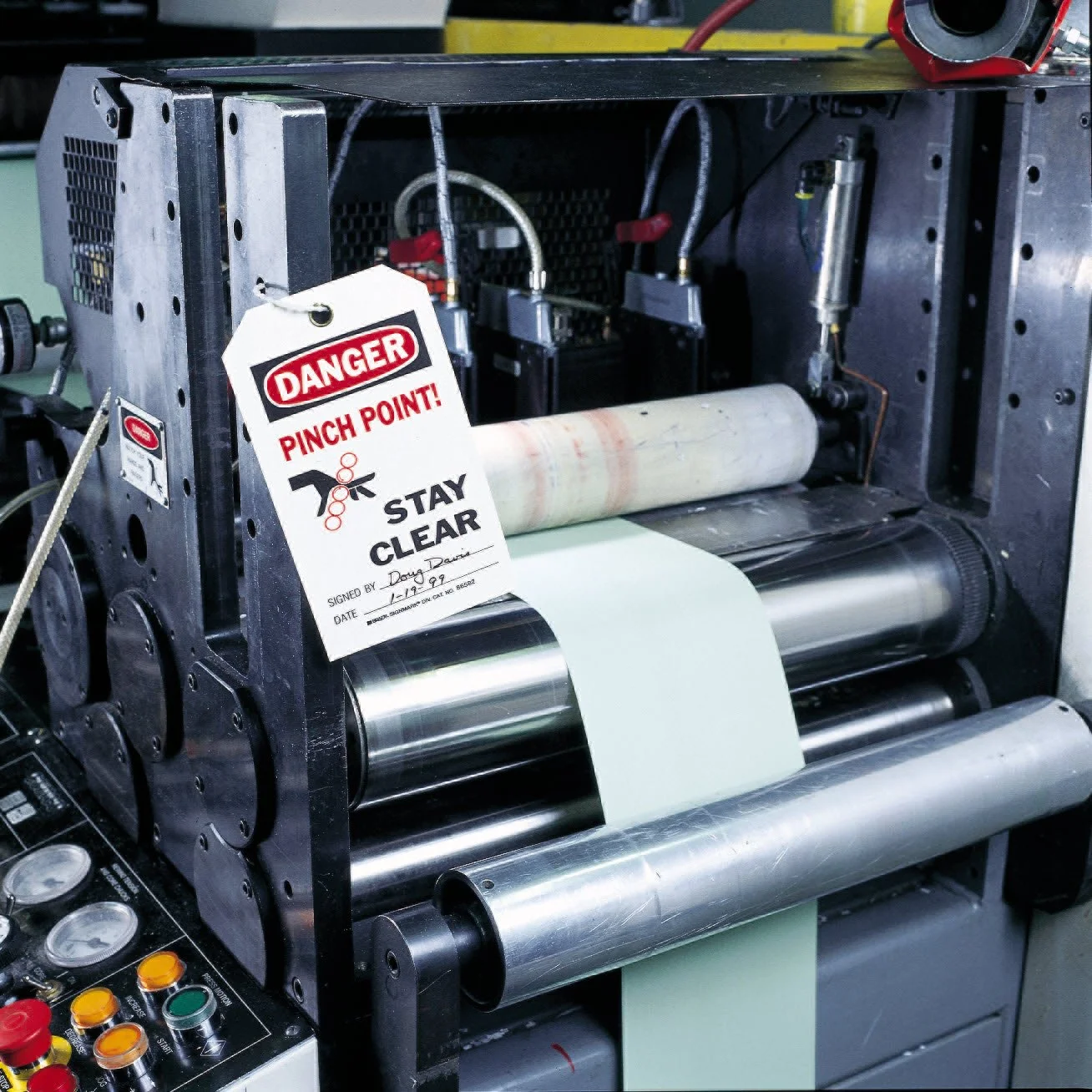 A close-up view of a printing machine with a warning tag that reads 'Danger Pinch Point! Stay Clear' attached to part of the machine. The machine is producing a sheet of light-colored paper or material.