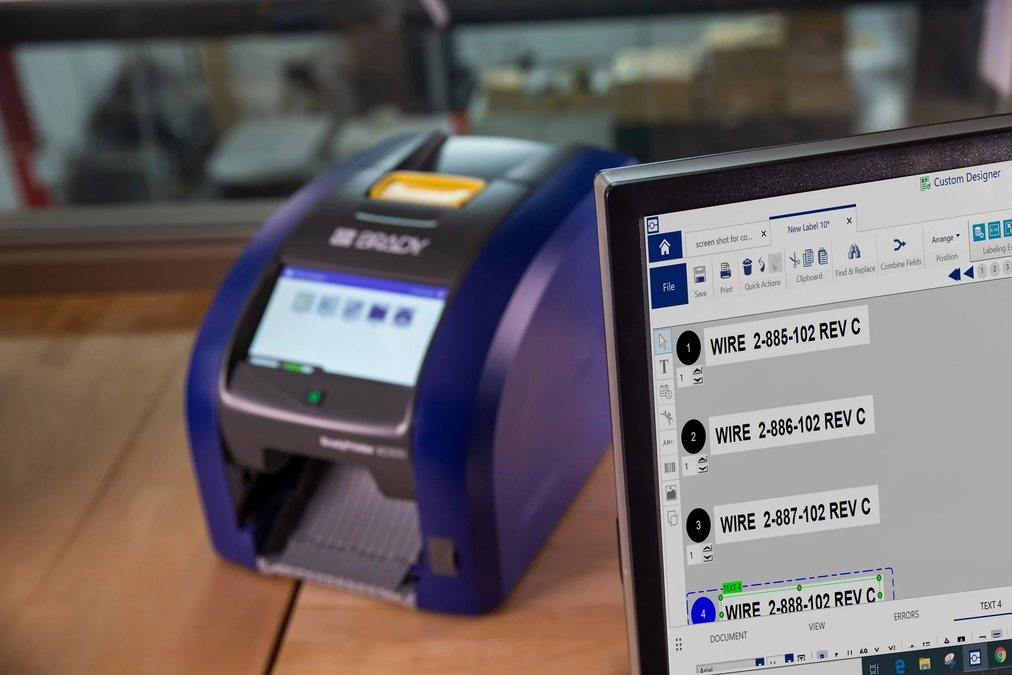 A computer screen displaying a design program with labels for wires, and a laser engraving machine in the background on a wooden table.