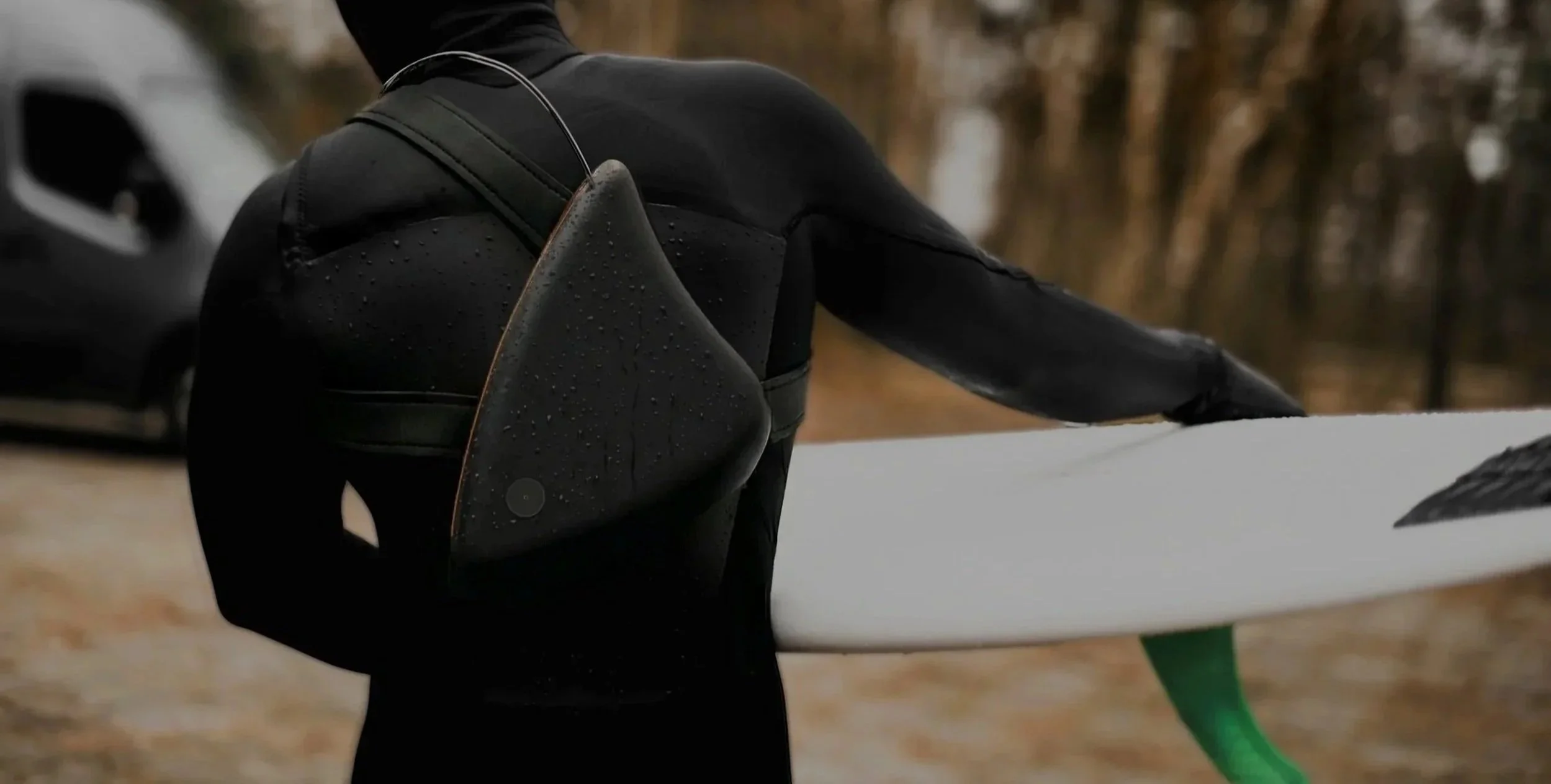 Person wearing black wetsuit holding a white surfboard with a green fin, standing outdoors with blurred trees in the background.