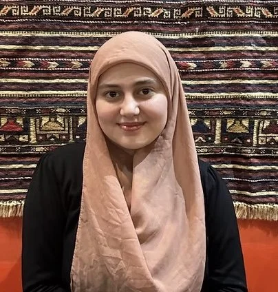 A woman wearing a beige hijab and black top, sitting in front of a multicolored patterned rug.