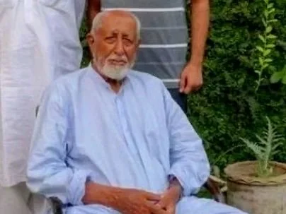 An elderly man with a white beard sitting outdoors, wearing a light blue kurta, surrounded by greenery.
