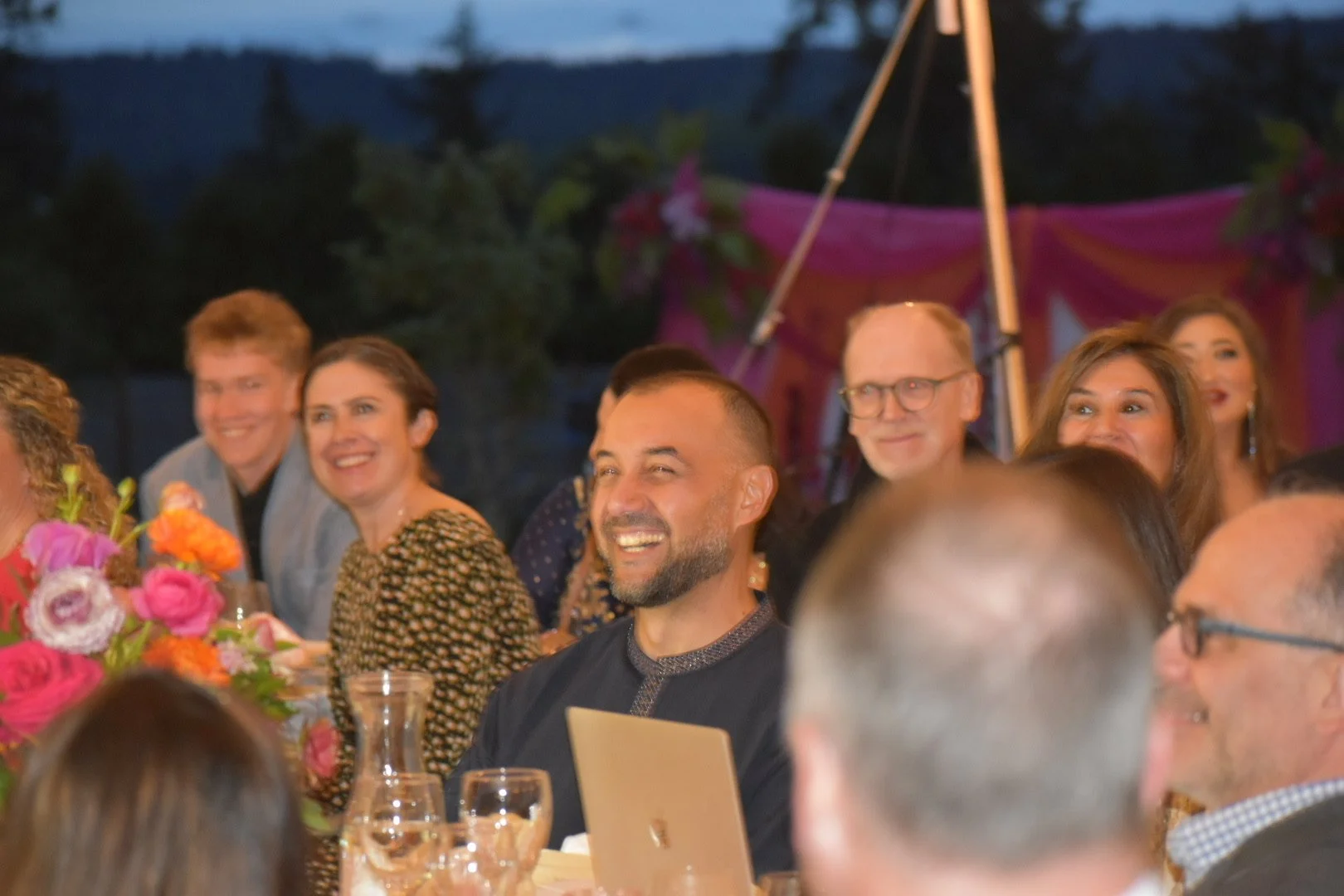 A group of people at a celebration or dinner, sitting around a table with flowers, smiling and laughing, in an outdoor setting during evening.