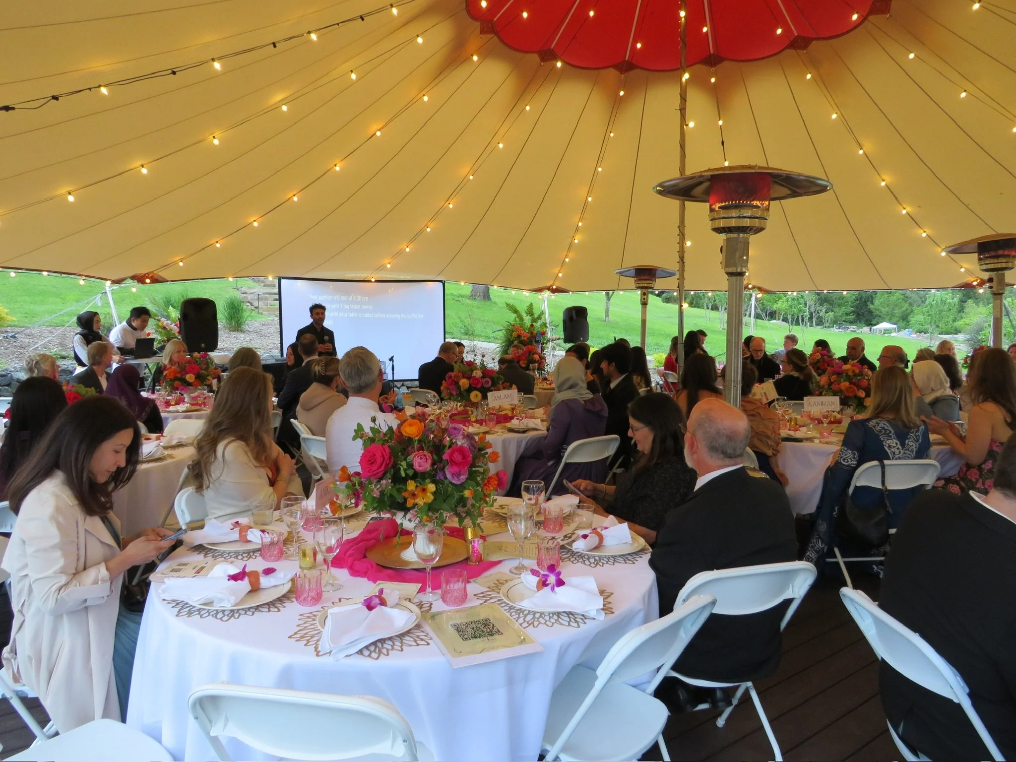 People attending a formal outdoor event under a large beige tent with string lights, seated at decorated round tables with floral centerpieces, while a speaker presents at the front.