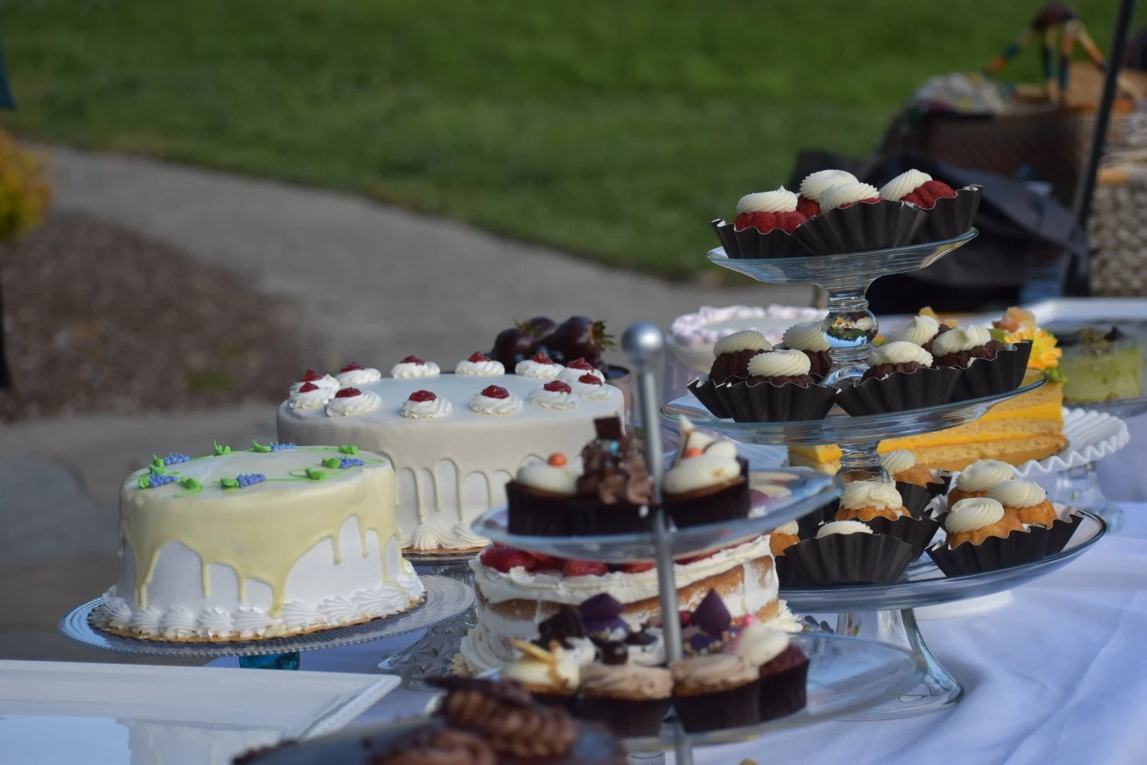 A dessert table with cakes and cupcakes for a celebration, outdoors on a white tablecloth.