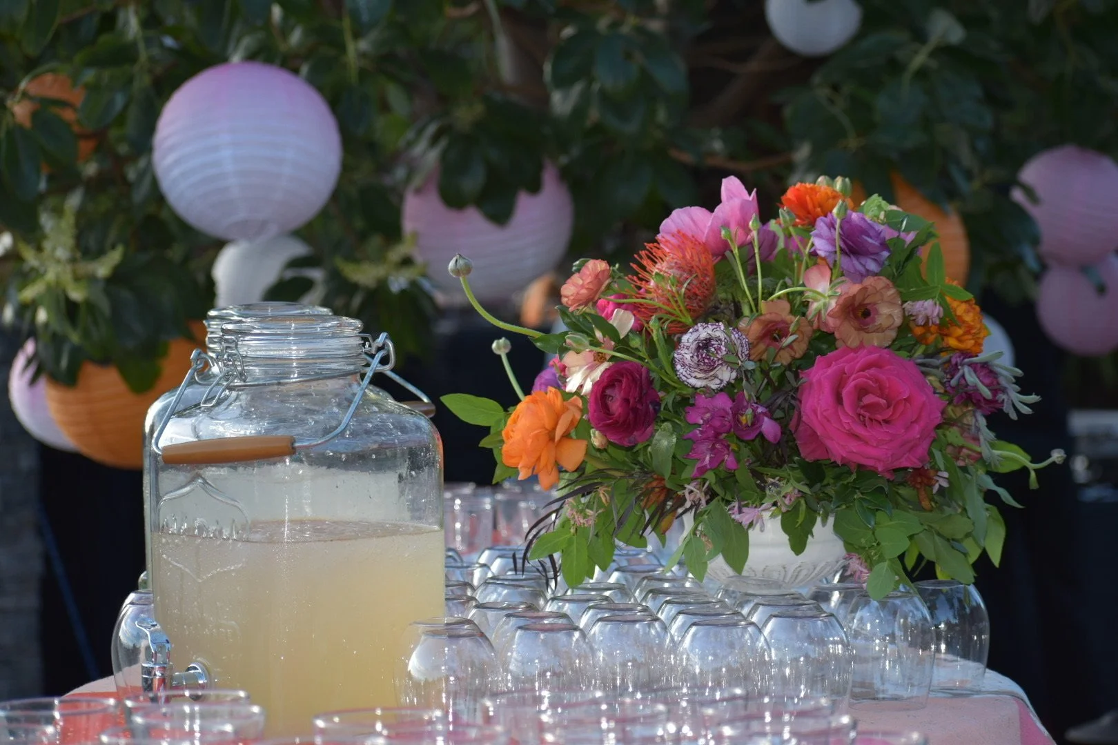 A table with a large bouquet of pink, orange, and purple flowers and a glass container filled with lemonade, with glass cups arranged in front. Lanterns and greenery are visible in the background.