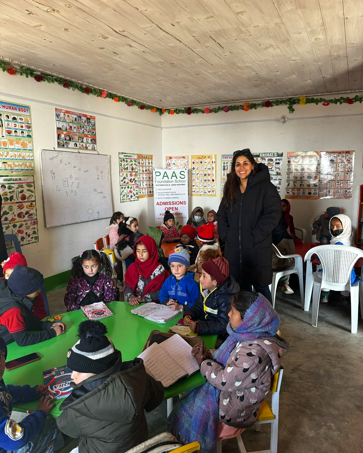 A classroom filled with young children sitting around green tables, some wearing winter clothes and hats, with a smiling woman standing among them. The classroom walls are decorated with educational posters about the human body and animals, and there is a sign for PAAS Foundation School.