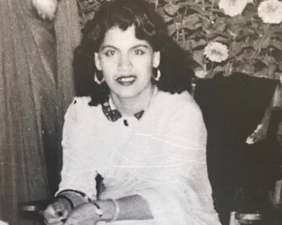 A black and white photograph of a woman with curly hair, wearing a white top, earrings, and lipstick, sitting at a table.