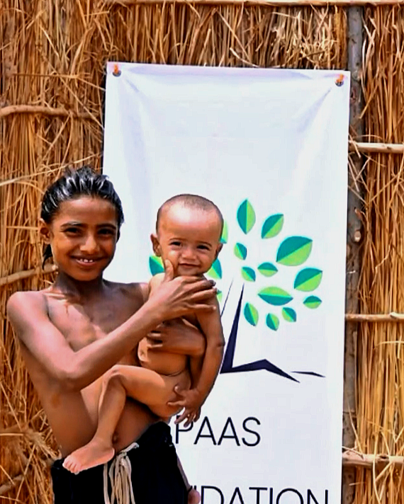 A young girl holding a smiling baby boy in front of a white banner with a green and blue tree logo and the text 'PAAS FOUNDATION', standing against a bamboo wall.