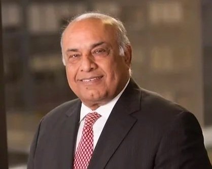 A portrait of an older man with gray hair, wearing a dark suit, white shirt, and red patterned tie, smiling in a professional setting.
