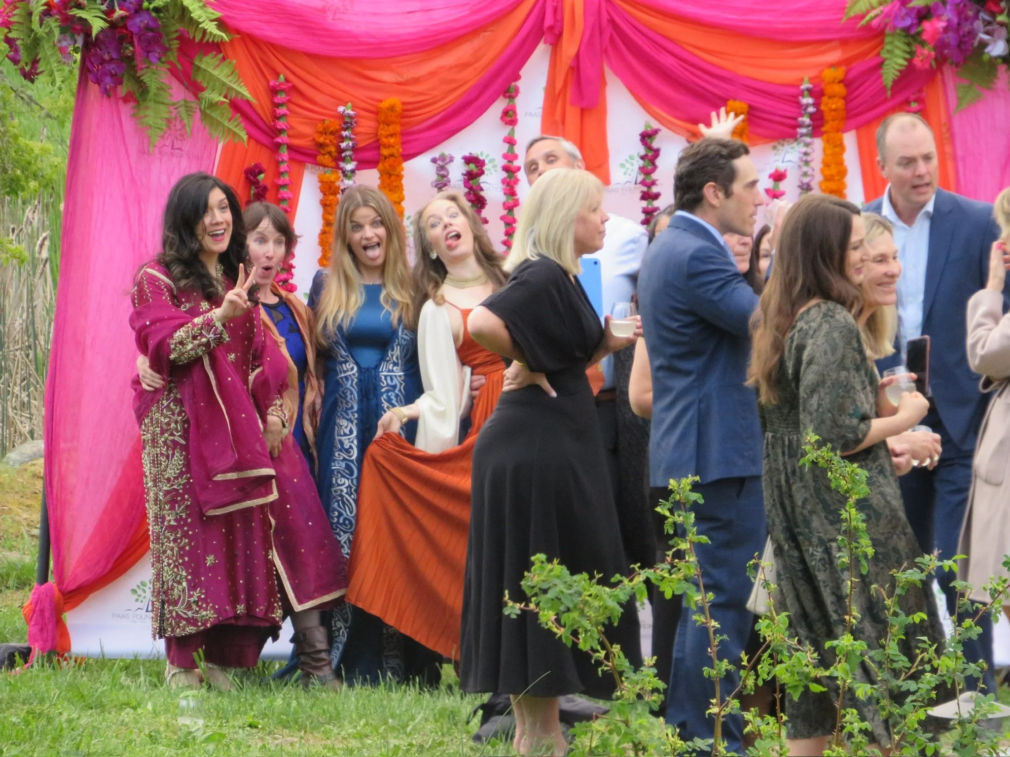 A group of people standing outdoors at a celebration or party, with some women dressed in traditional South Asian attire, in front of a colorful decorated backdrop with pink, orange, and purple fabric and flower garlands.