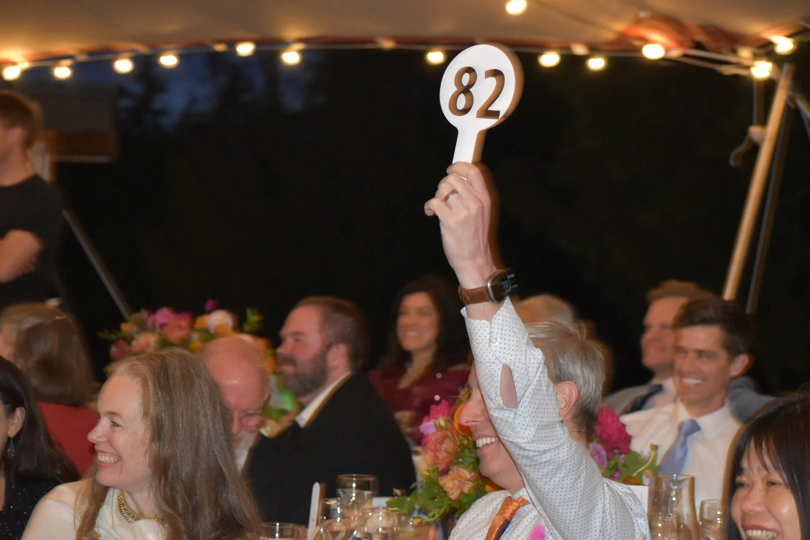 A man smiling and holding up a card with the number 82 at a formal event or auction, surrounded by other smiling attendees sitting at a table adorned with flowers.