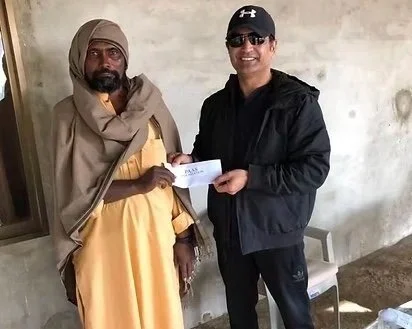 Two men standing indoors, one dressed in traditional clothing and the other in black athletic wear, exchanging a check.