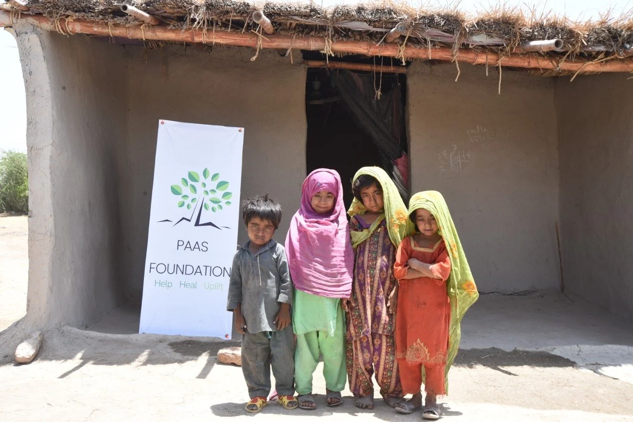 Four children standing in front of a simple mud house with a PAAS Foundation banner nearby. The children are dressed in colorful traditional clothing and head coverings.