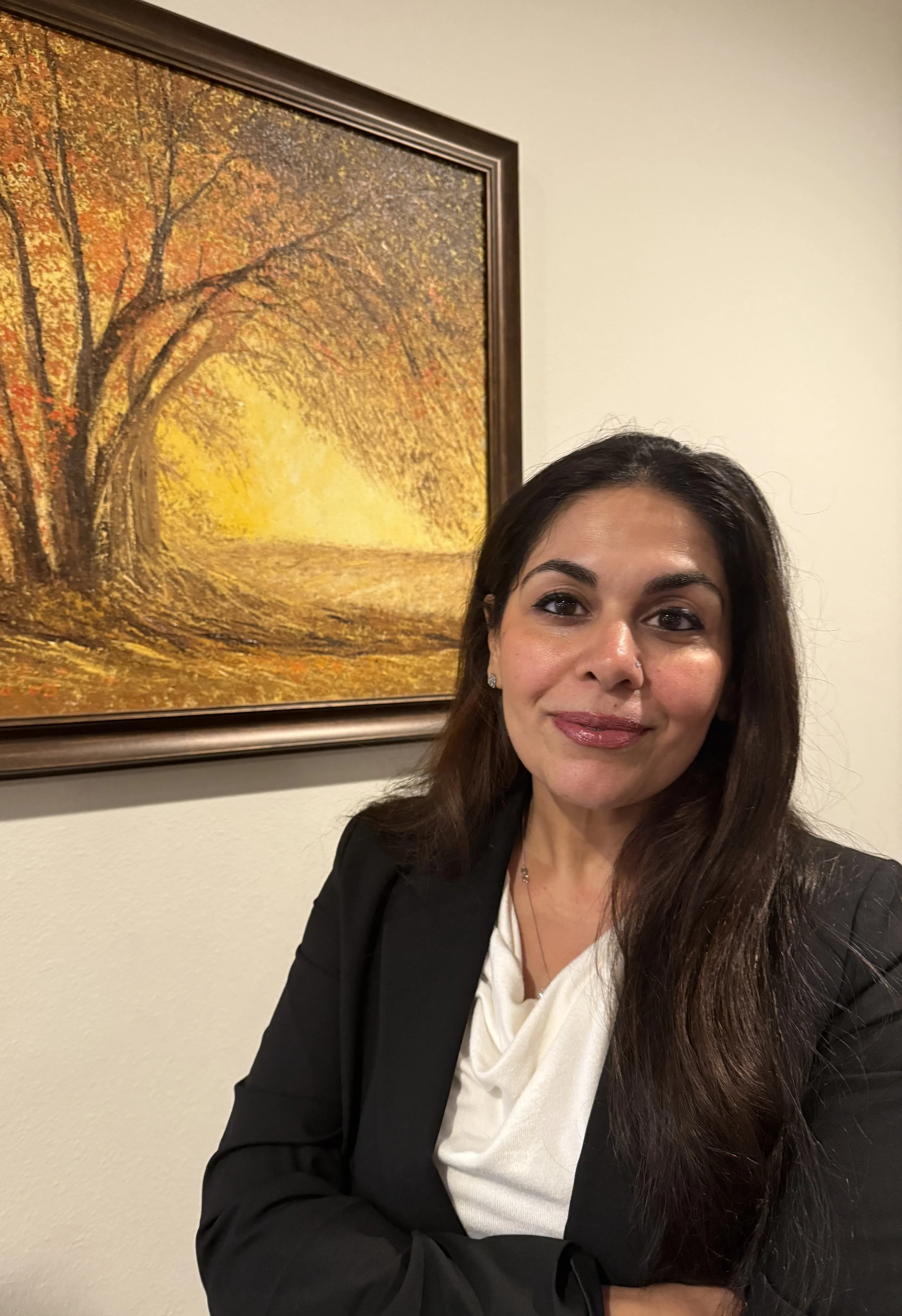 A woman with long dark hair wearing a black blazer and a white blouse, standing in front of a painting of a tree with autumn leaves and a bright yellow setting sun.