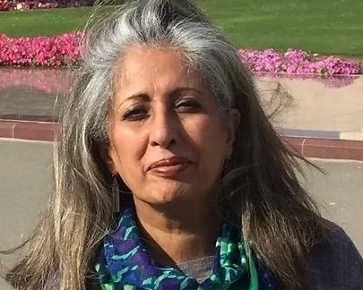 A woman with gray hair wearing a blue patterned shirt stands outdoors with pink flowers and a green lawn in the background.