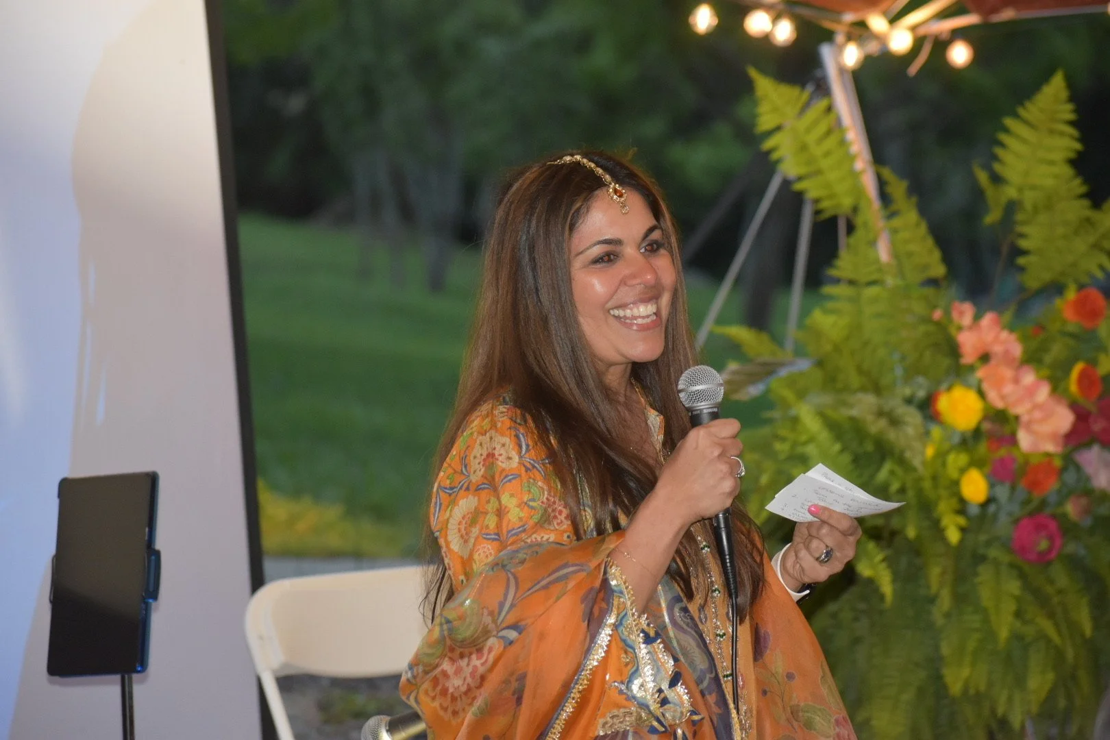 A woman dressed in a colorful traditional outfit holding a microphone and reading from a paper, smiling during an outdoor event with greenery and flowers in the background.