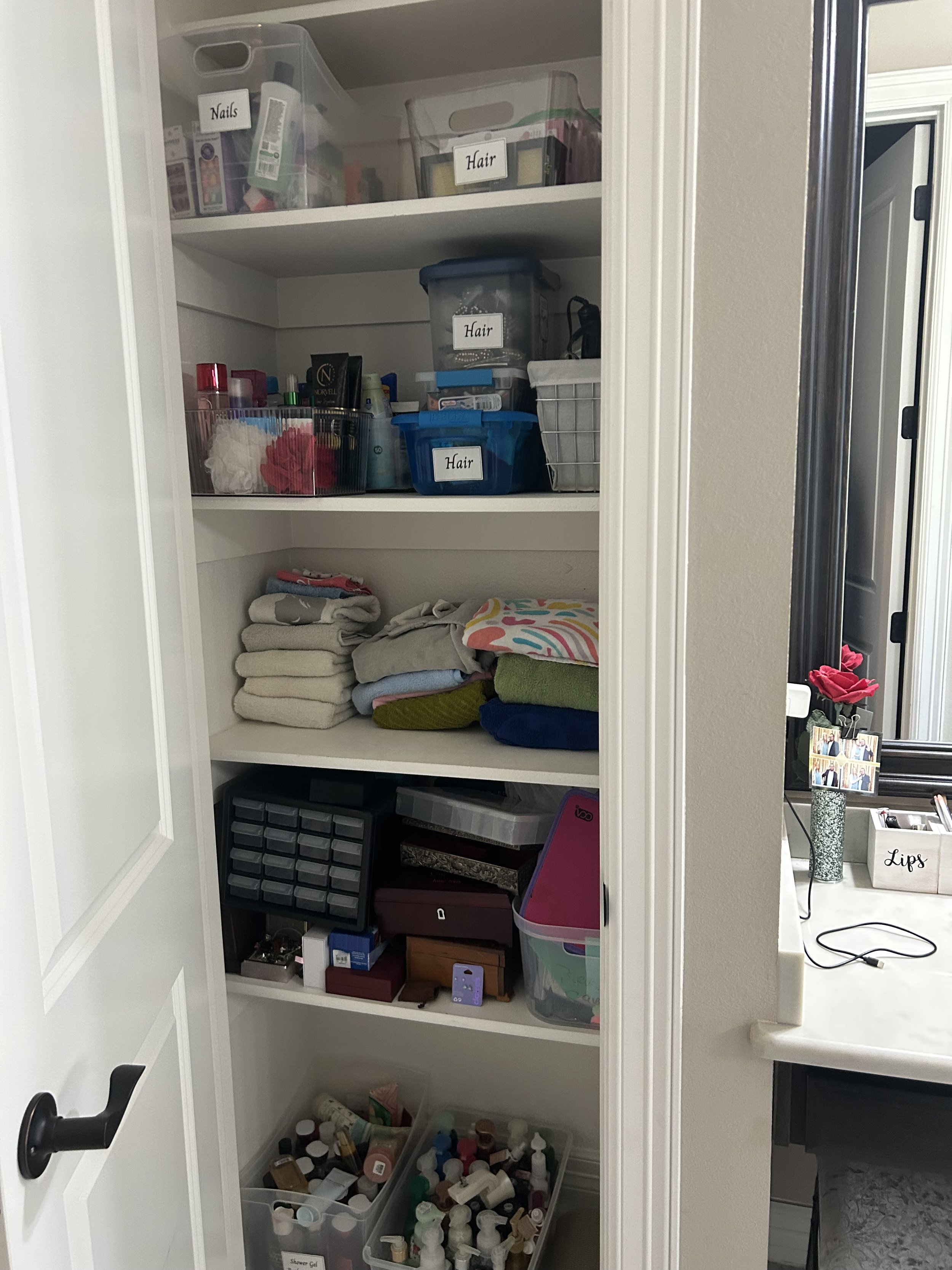 A closet with labeled shelves containing toiletries, towels, and hair supplies. The top shelf has labeled containers for nails and hair. The middle shelves hold hair products, towels, and organizational boxes. The bottom has bottles and toiletries.