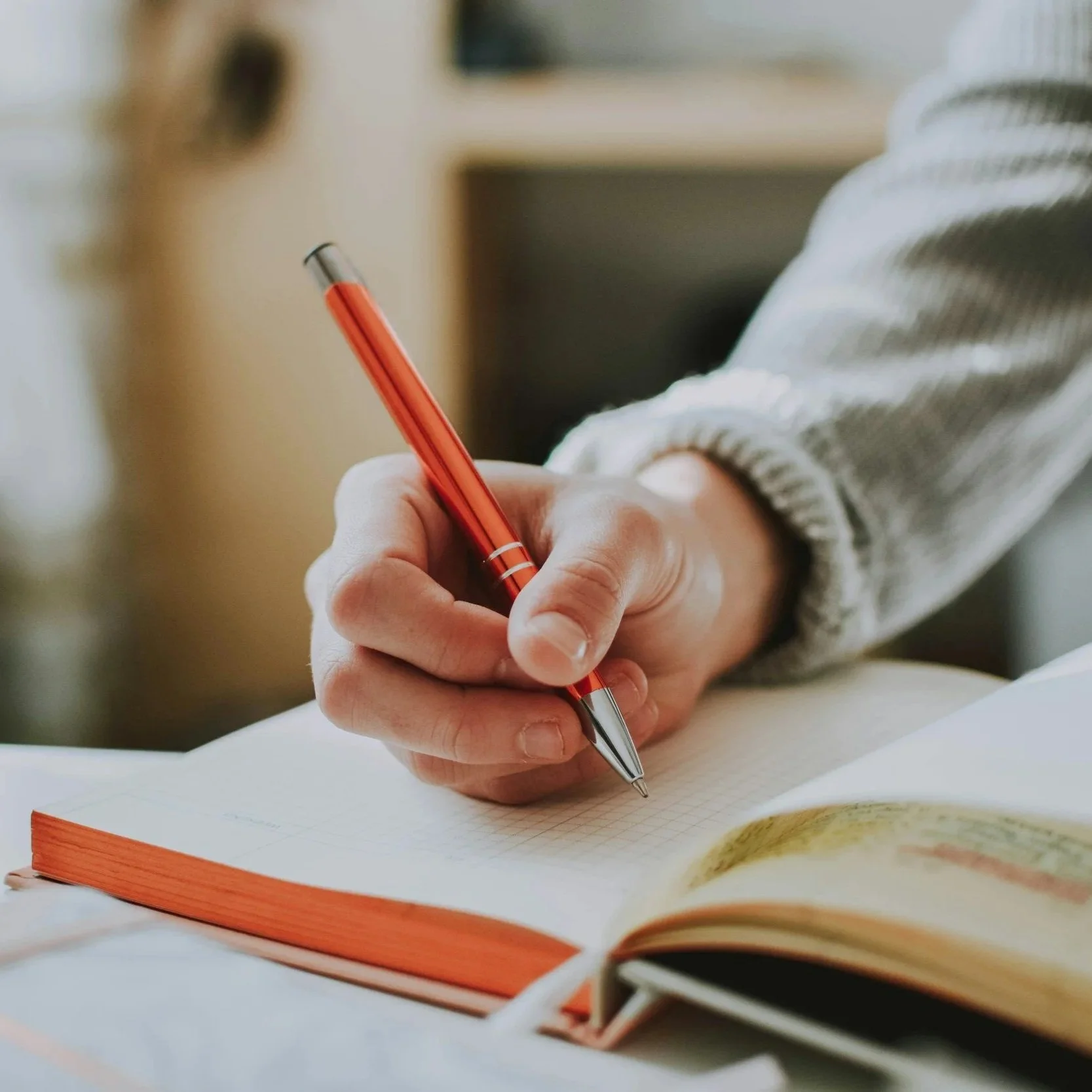 A person writing in a notebook with a red and silver pen, wearing a gray sweater.