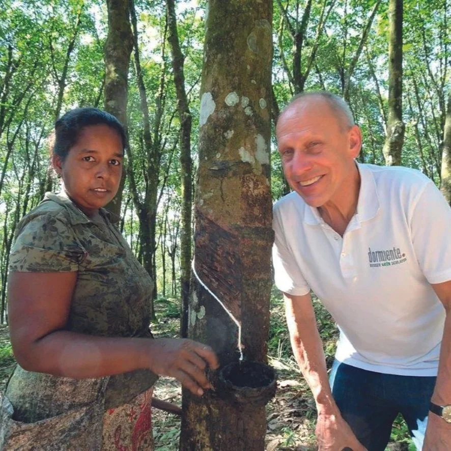 A woman and a man outdoors in a forested area with tall trees, planting a tree together and putting sap, with the woman holding a small container and the man smiling.