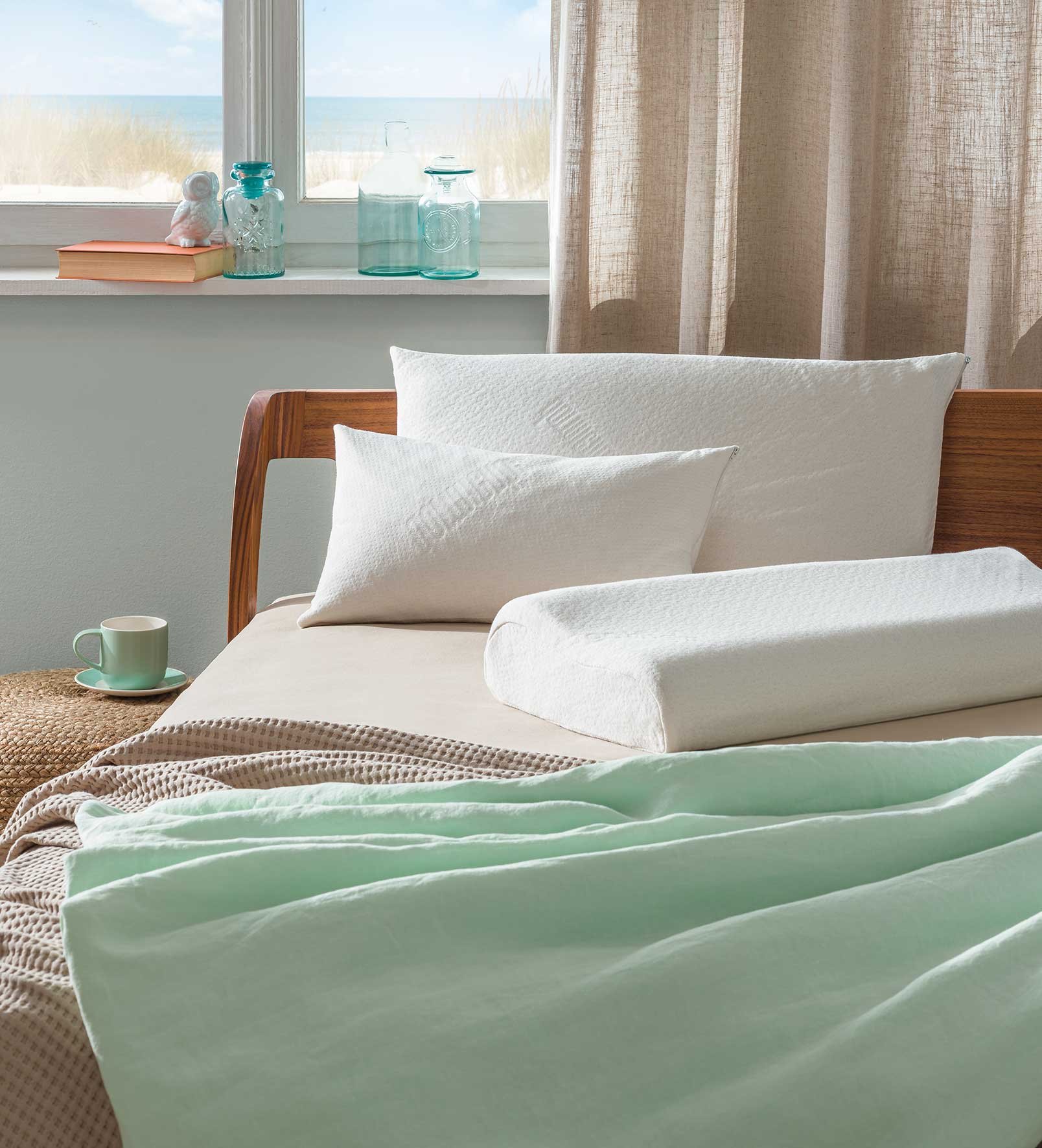 A bedroom with two pillows and a bolster on a bed with pastel-colored bedding, a window with a view of the beach, and a window sill with glass jars, a book, a cup, and a saucer.