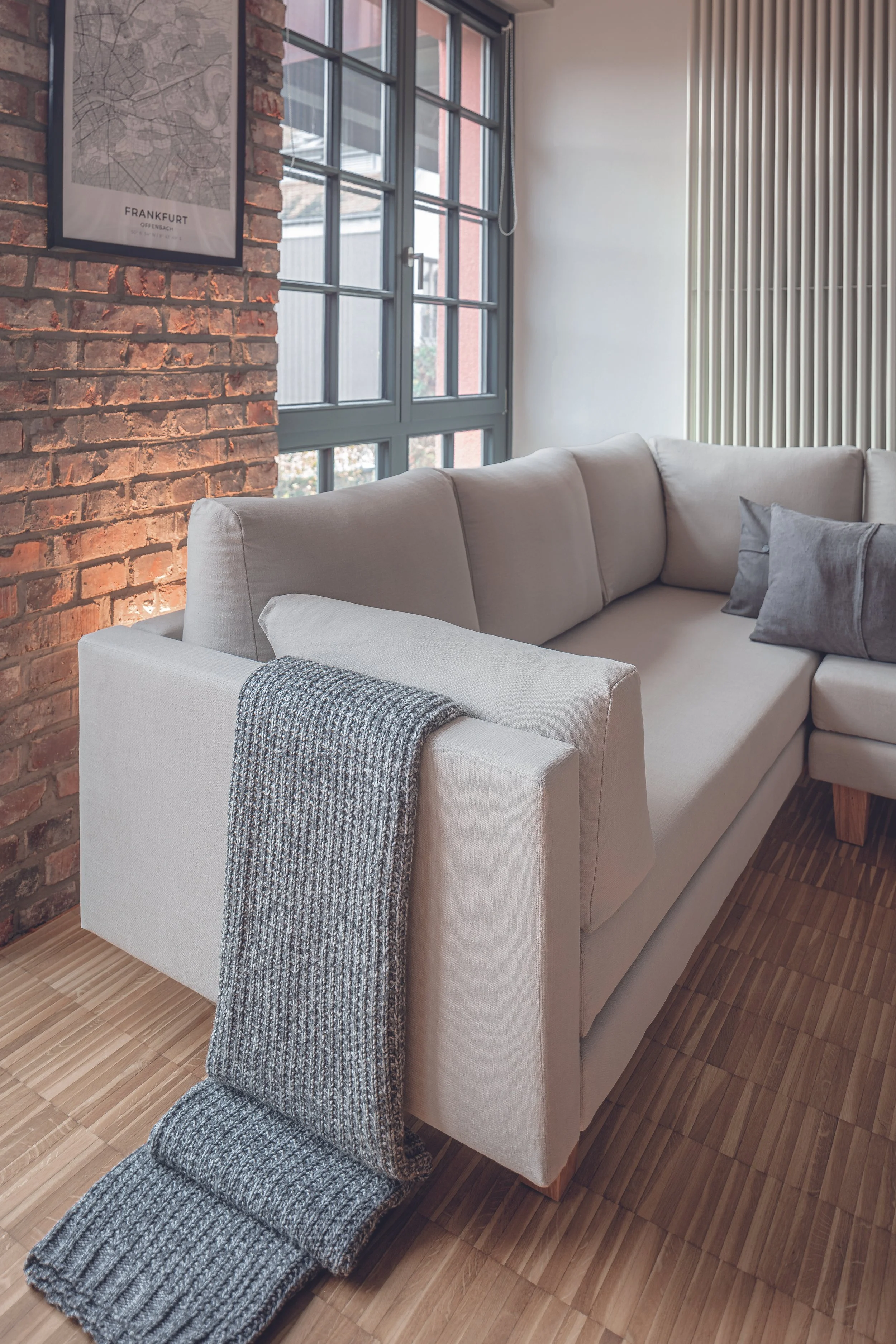 Modern living room with a beige sectional sofa, gray knitted throw blanket, brick accent wall, large windows, and minimal decor.