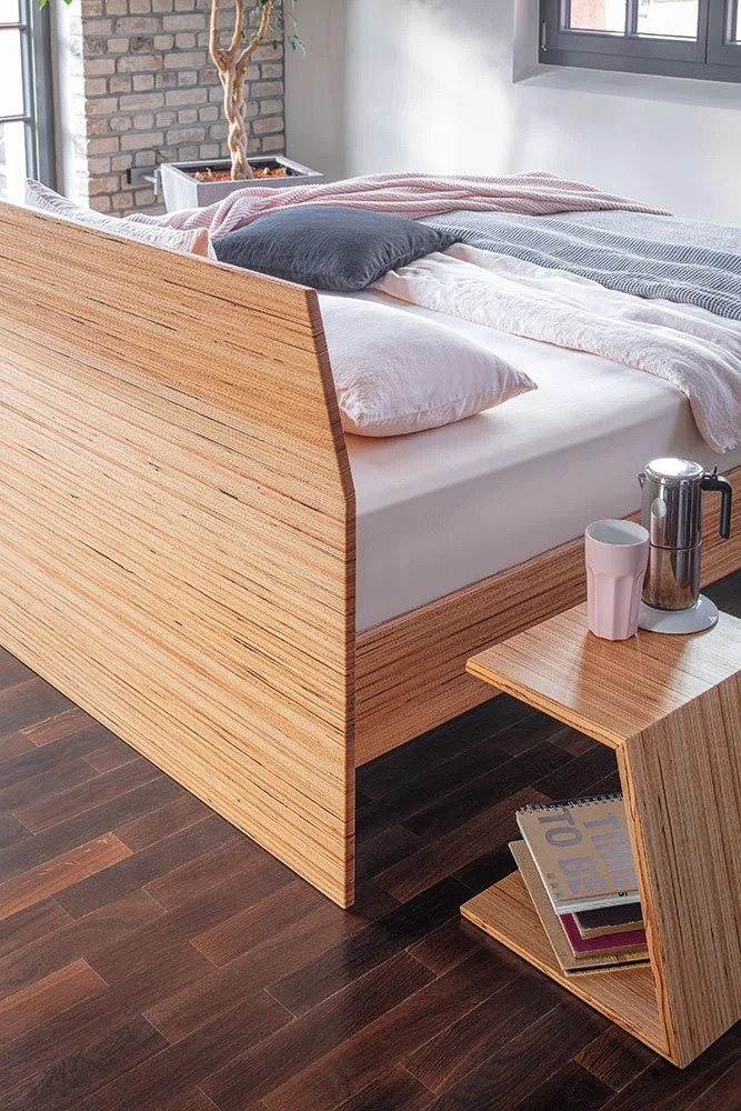 Modern bedroom with wooden bed frame, side table, books, coffee pot, and cups. Features dark hardwood floor, brick wall, large window, and potted plant in corner.