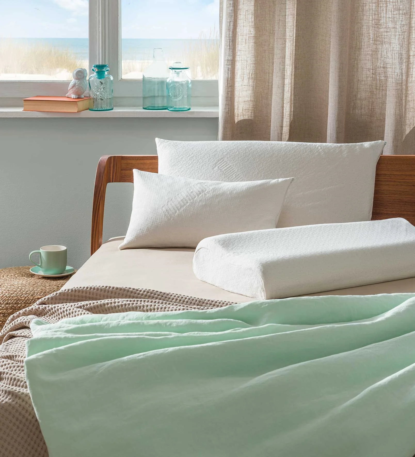 Cozy bedroom scene with a bed featuring white pillows and light mint green bedding, a wooden headboard, a cup on a woven stool, and glass bottles on a window sill with an ocean view.