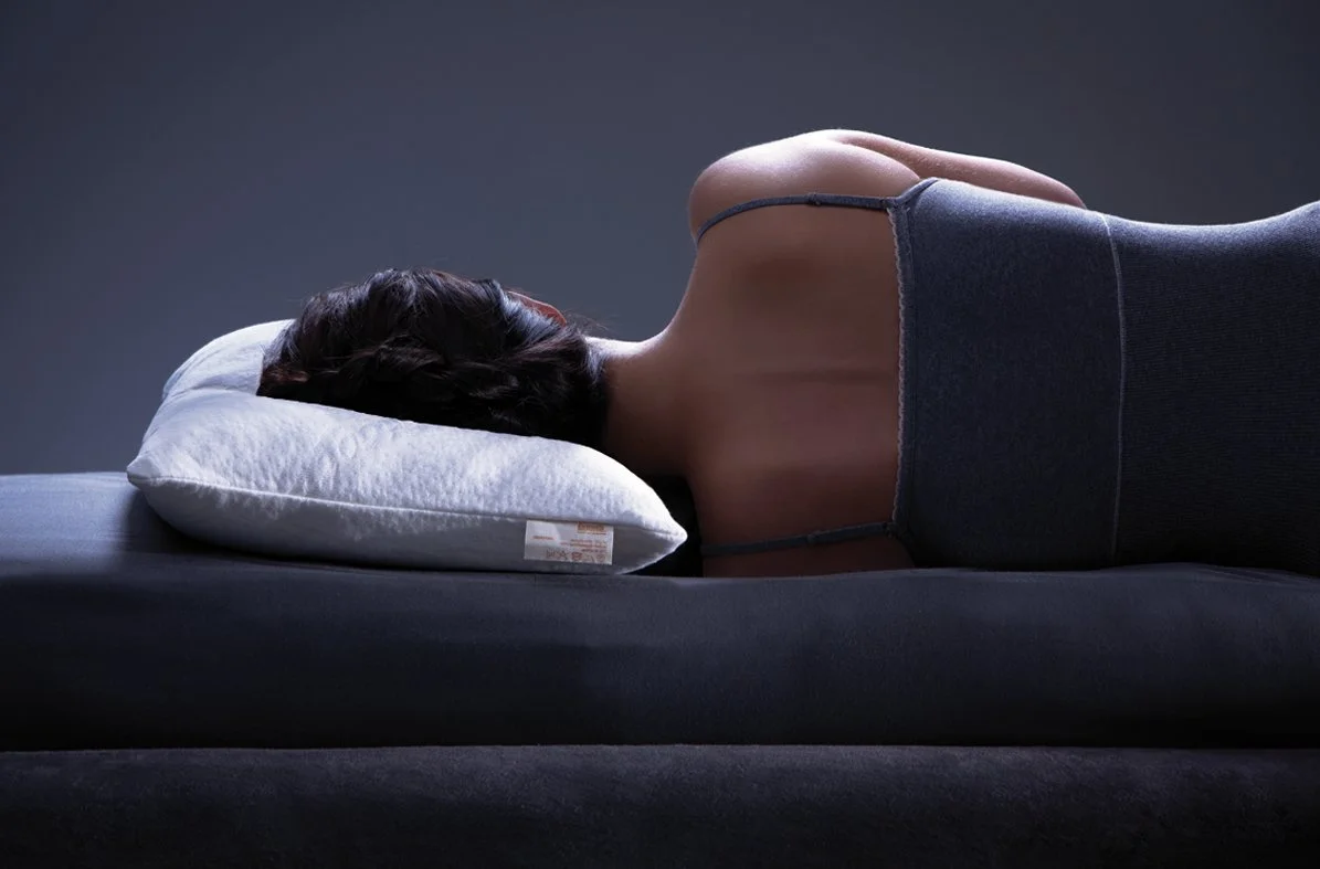 Person sleeping on their side with a pillow on a bed, wearing a tank top, in a dimly lit room.