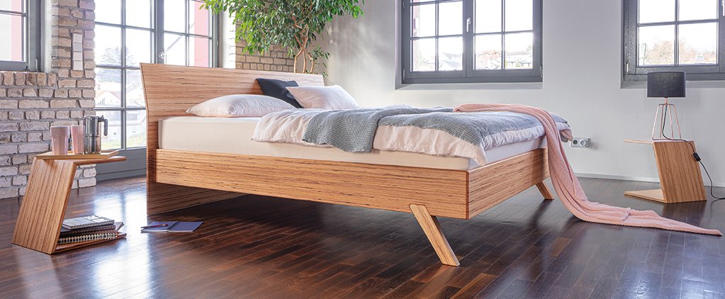 Modern bedroom with a wooden bed frame, gray blanket, and pink throw; side tables with books and a lamp; large windows and a potted plant.