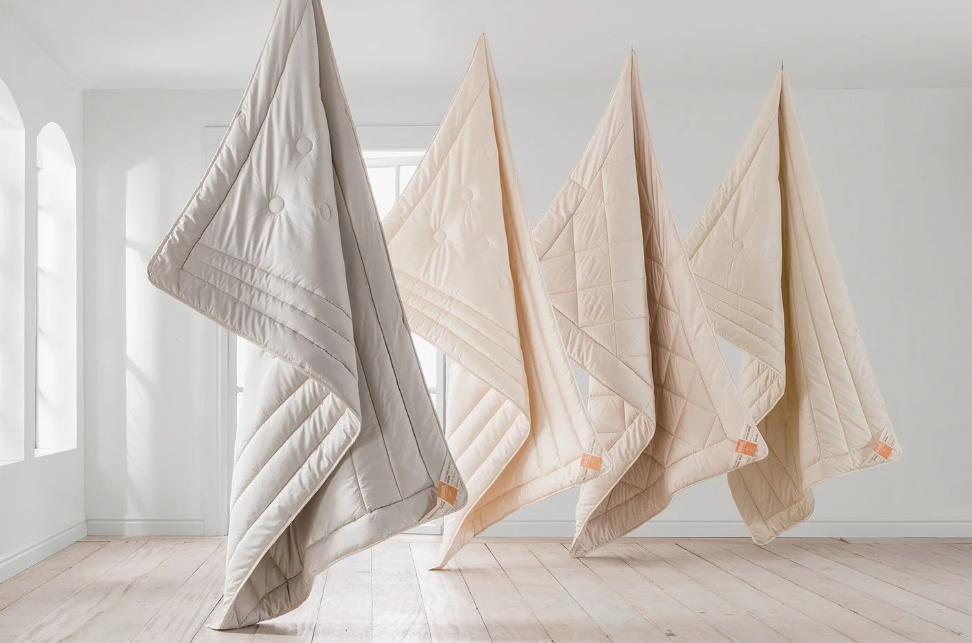 Four beige and cream-colored quilted duvets hanging in a bright room.