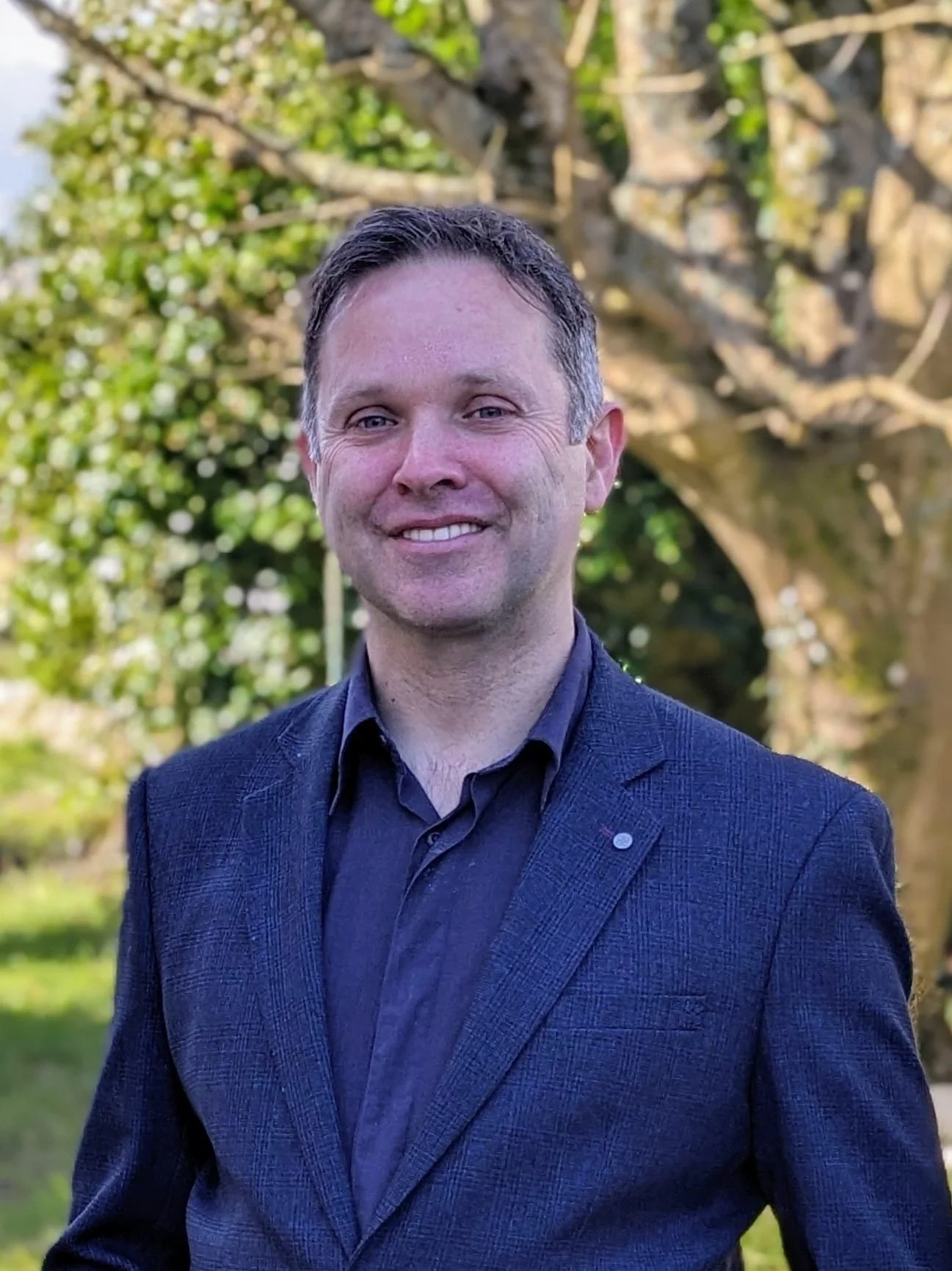Man in dark suit smiling, standing outdoors with green foliage and tree in the background.