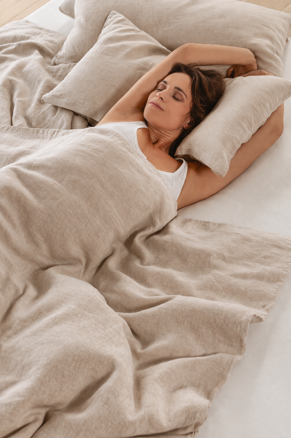 A woman sleeping peacefully on a bed with beige linen sheets and pillows, wearing a white tank top.