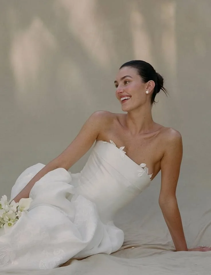 A woman in a white wedding dress sitting on a beach with a smile, holding a bouquet of white flowers.