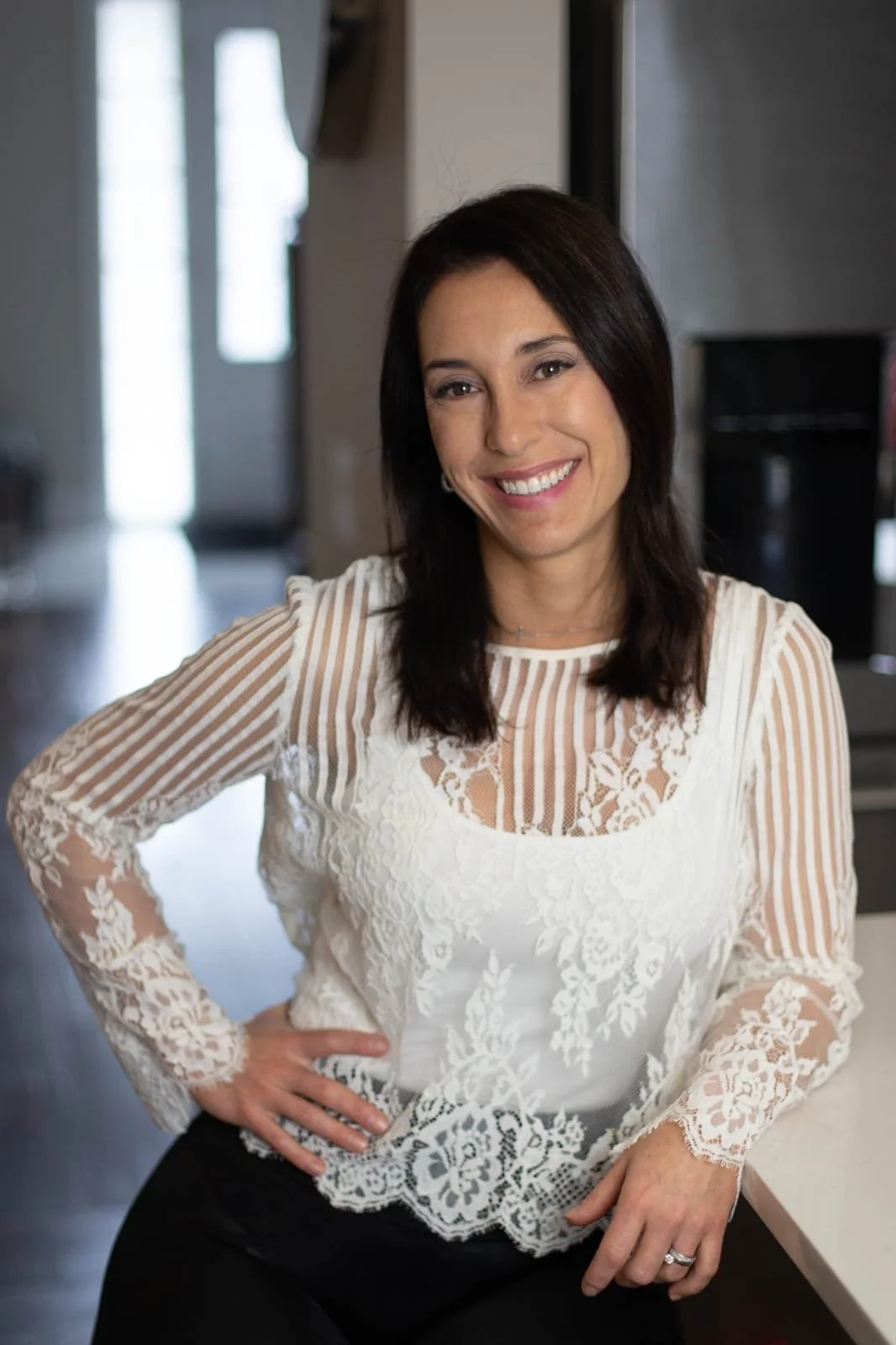 A woman with dark hair smiling, wearing a white lace and striped sheer top, sitting in a kitchen or dining area.