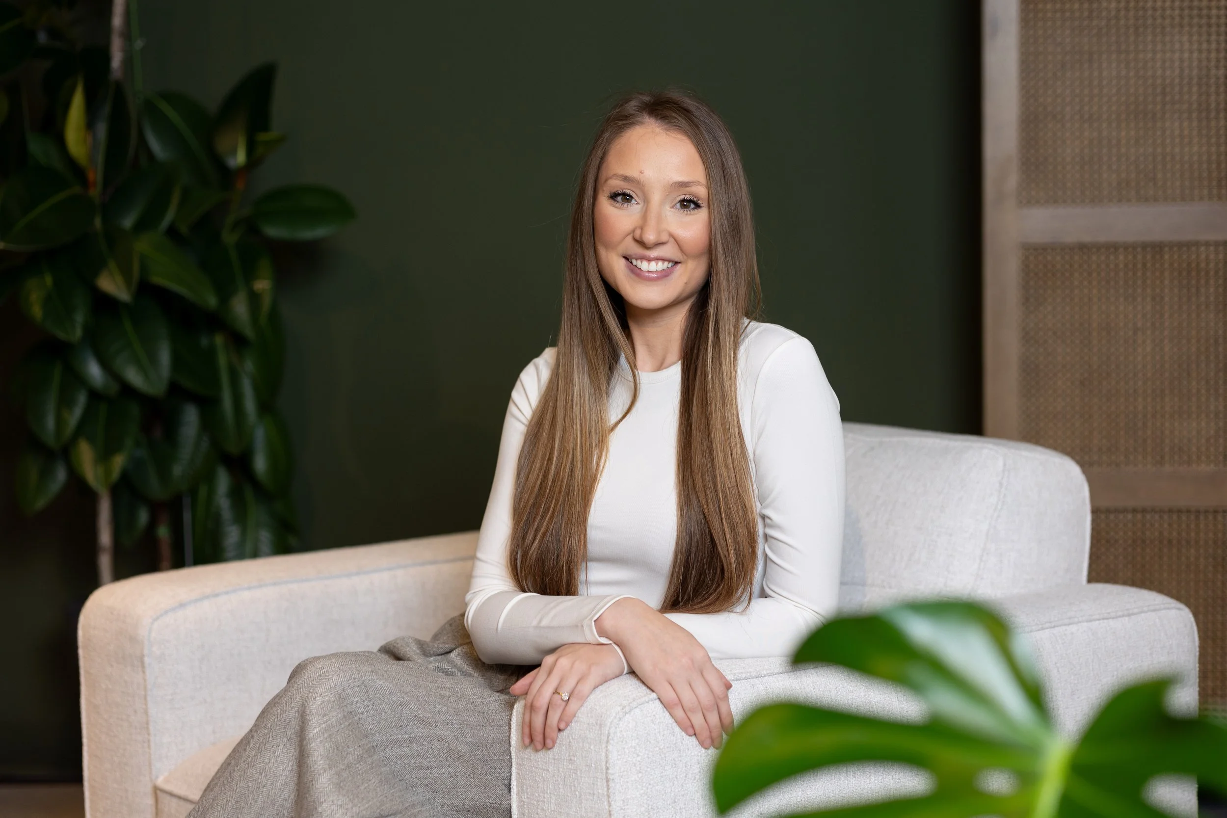A young woman with long brown hair and a white long-sleeve shirt sitting on a beige sofa, smiling at the camera, with dark green wall and a porcupine plant in the background.