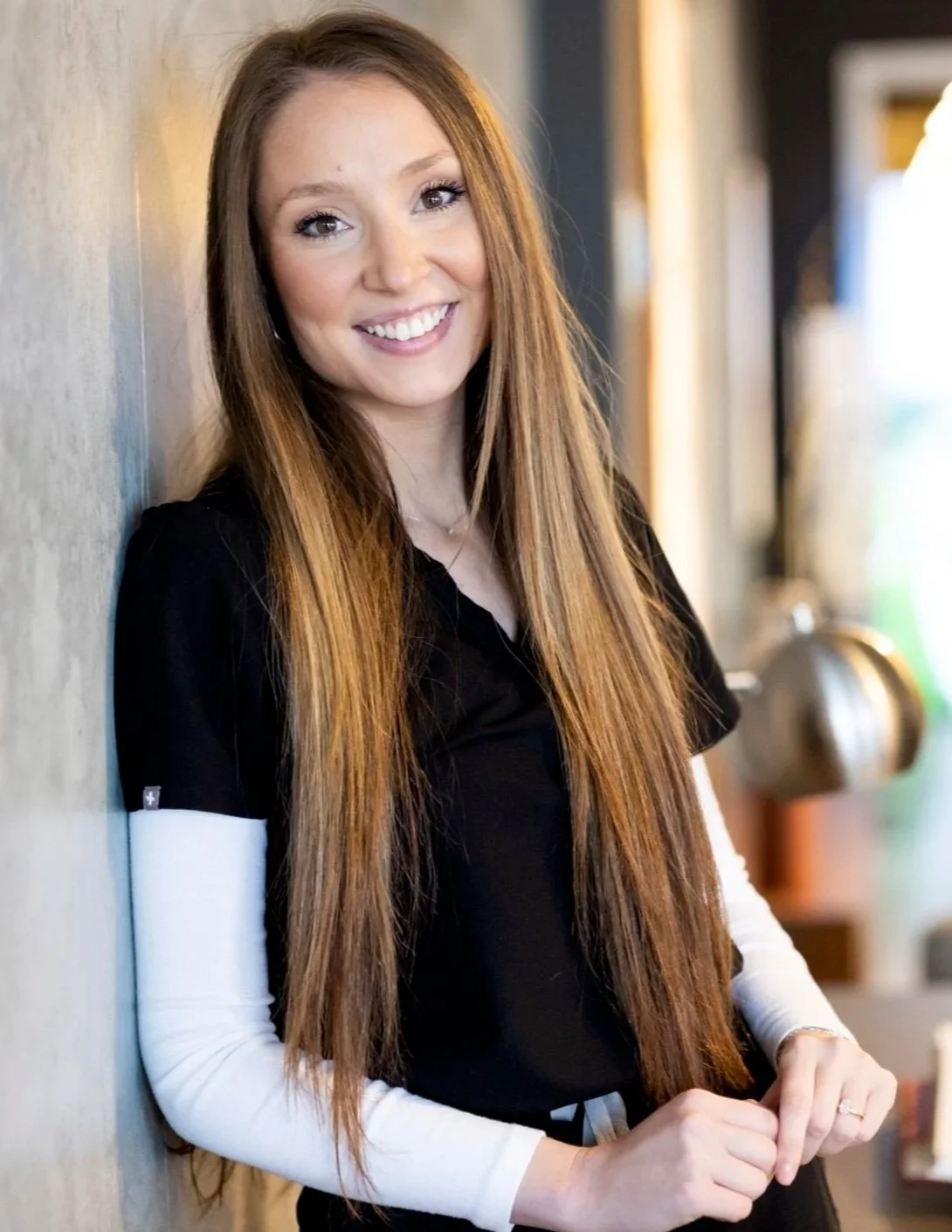 A young woman with long red hair smiling, standing against a wall, wearing a black top with white sleeves, in a casual indoor setting.