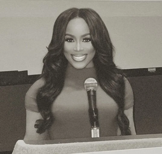 A woman with long wavy hair smiling at the camera, sitting behind a podium with a microphone.