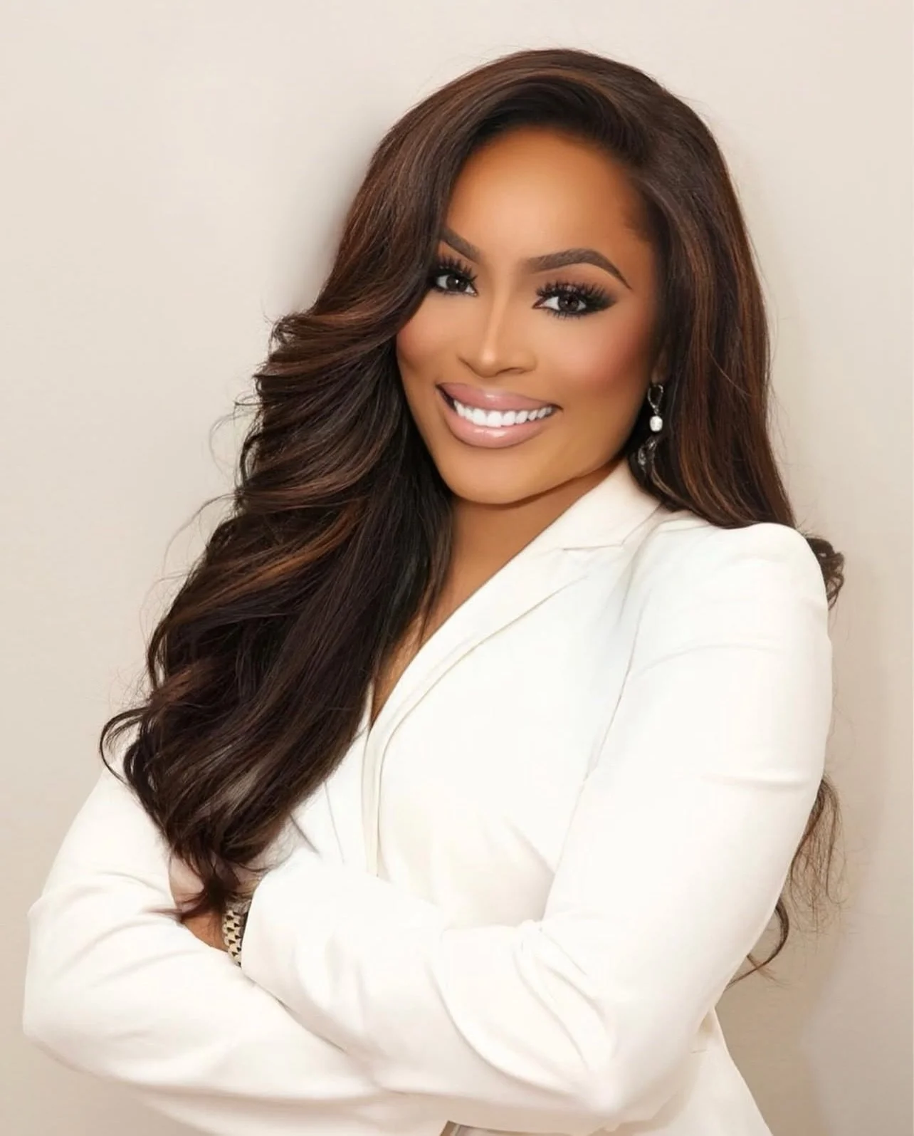 A smiling woman with long, dark brown hair styled in waves, wearing a white blazer and pearl earrings.