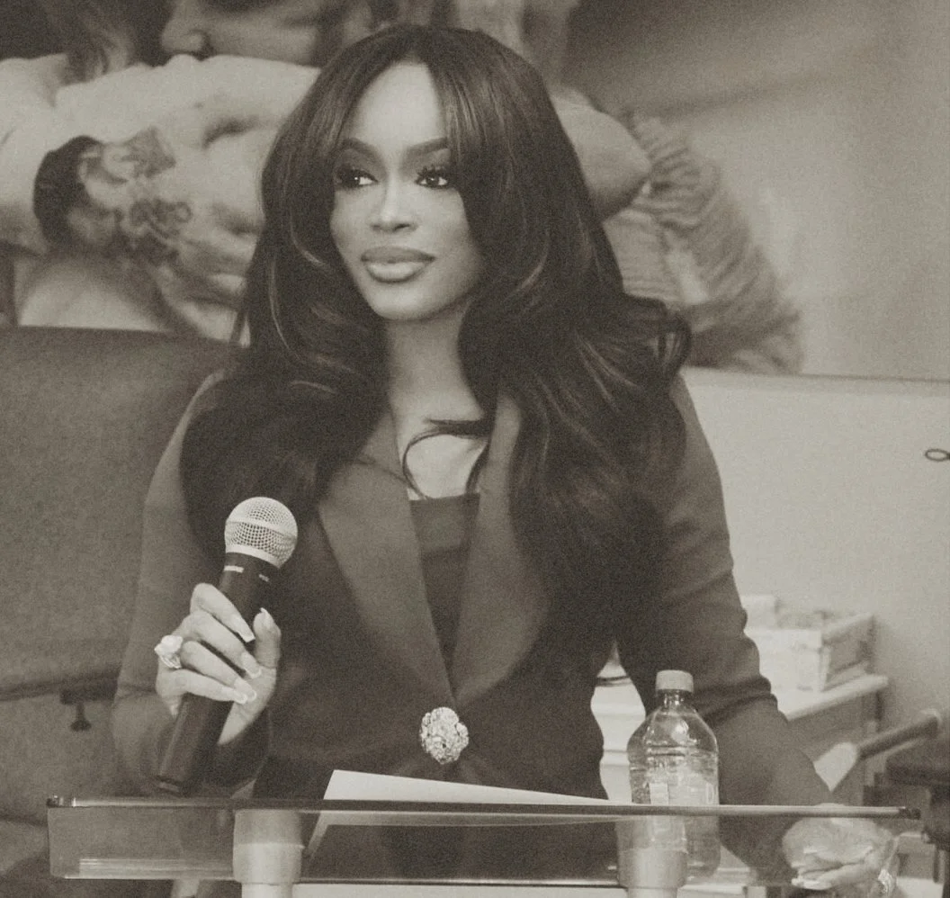 A woman speaking into a microphone at a formal event, sitting at a table with a water bottle and papers, with a large painting in the background.