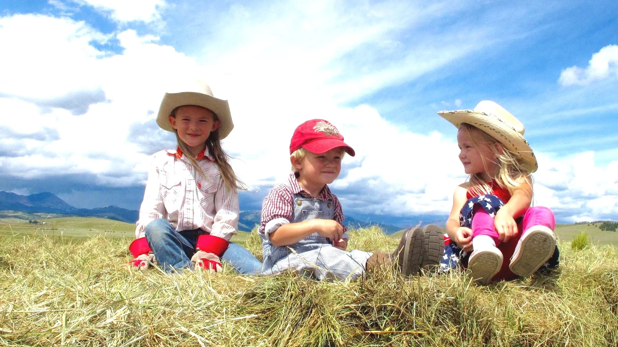 Three children sitting on a grassy field under a partly cloudy sky, wearing cowboy hats and casual clothing, with mountains in the background.