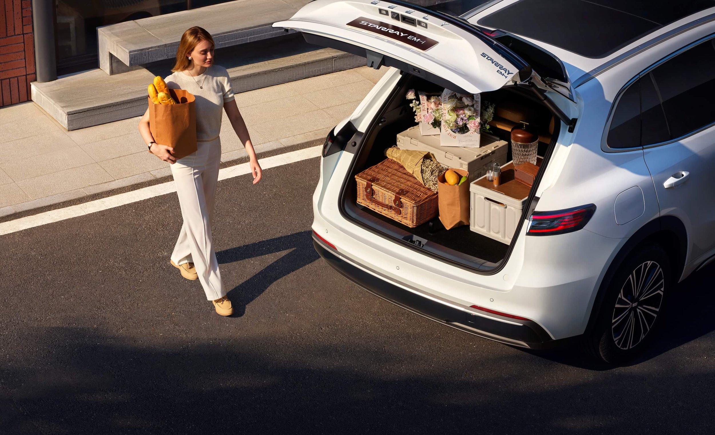 Mujer con blusa blanca y pantalones blancos cargando una bolsón de compras con pan, junto a un auto con maletero abierto lleno de cajas, flores y compras.