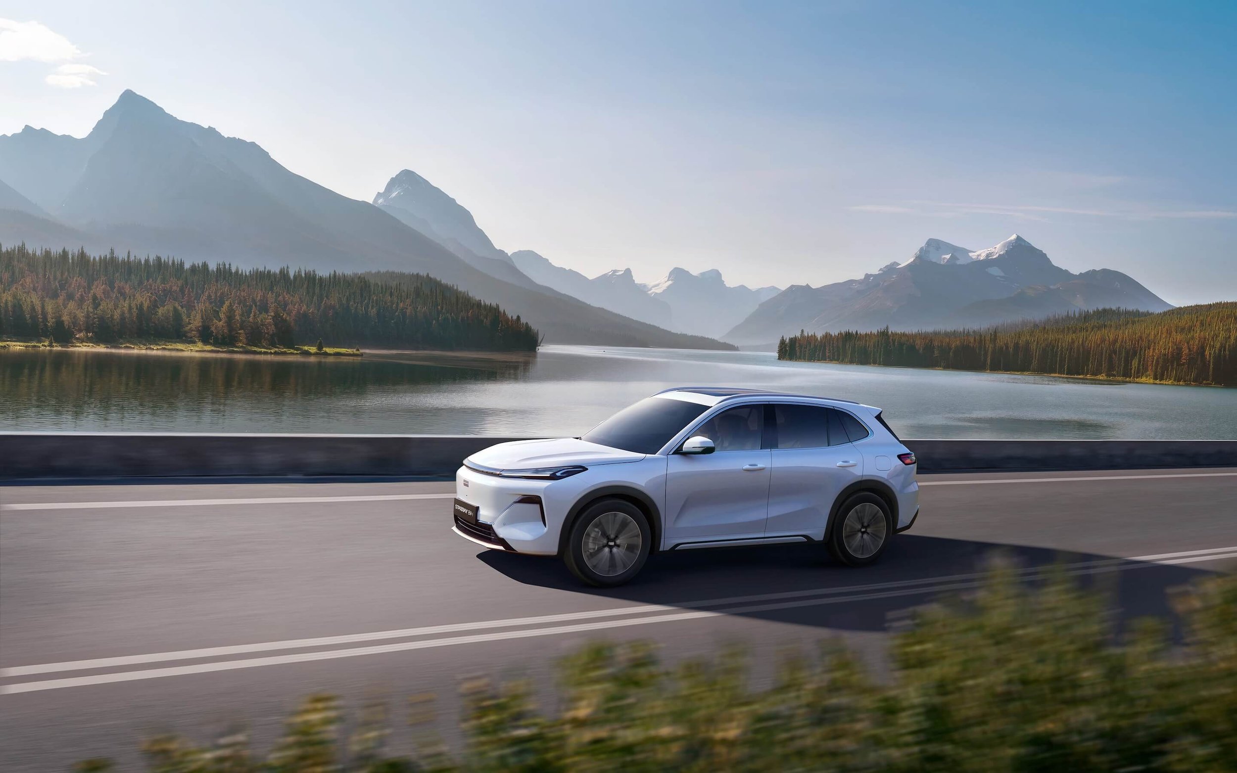 Auto eléctrico plateado conduciendo por una carretera junto a un lago con montañas y bosque de fondo