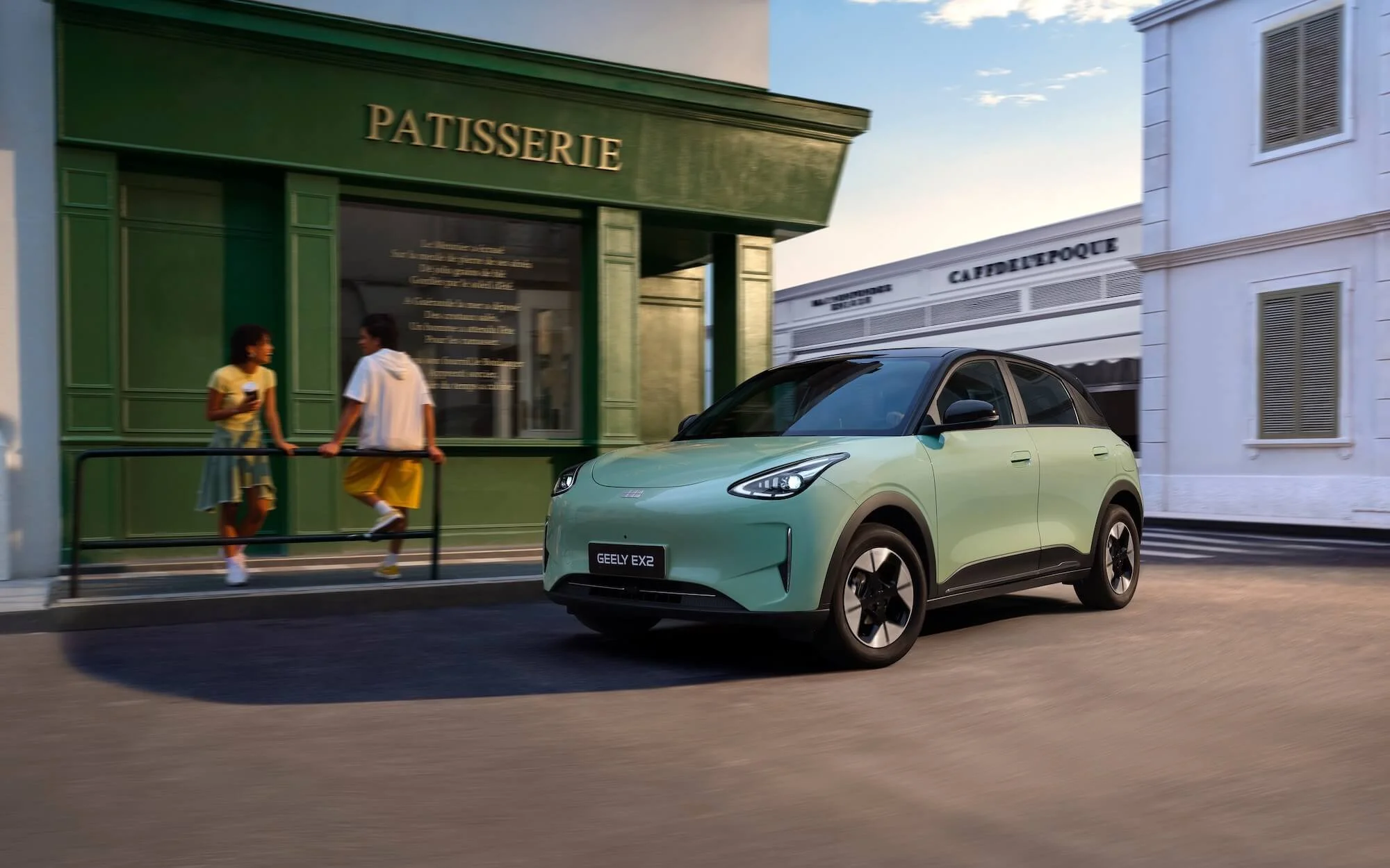 Coche eléctrico verde claro circulando por la ciudad con dos personas jóvenes conversando frente a una tienda de repostería.