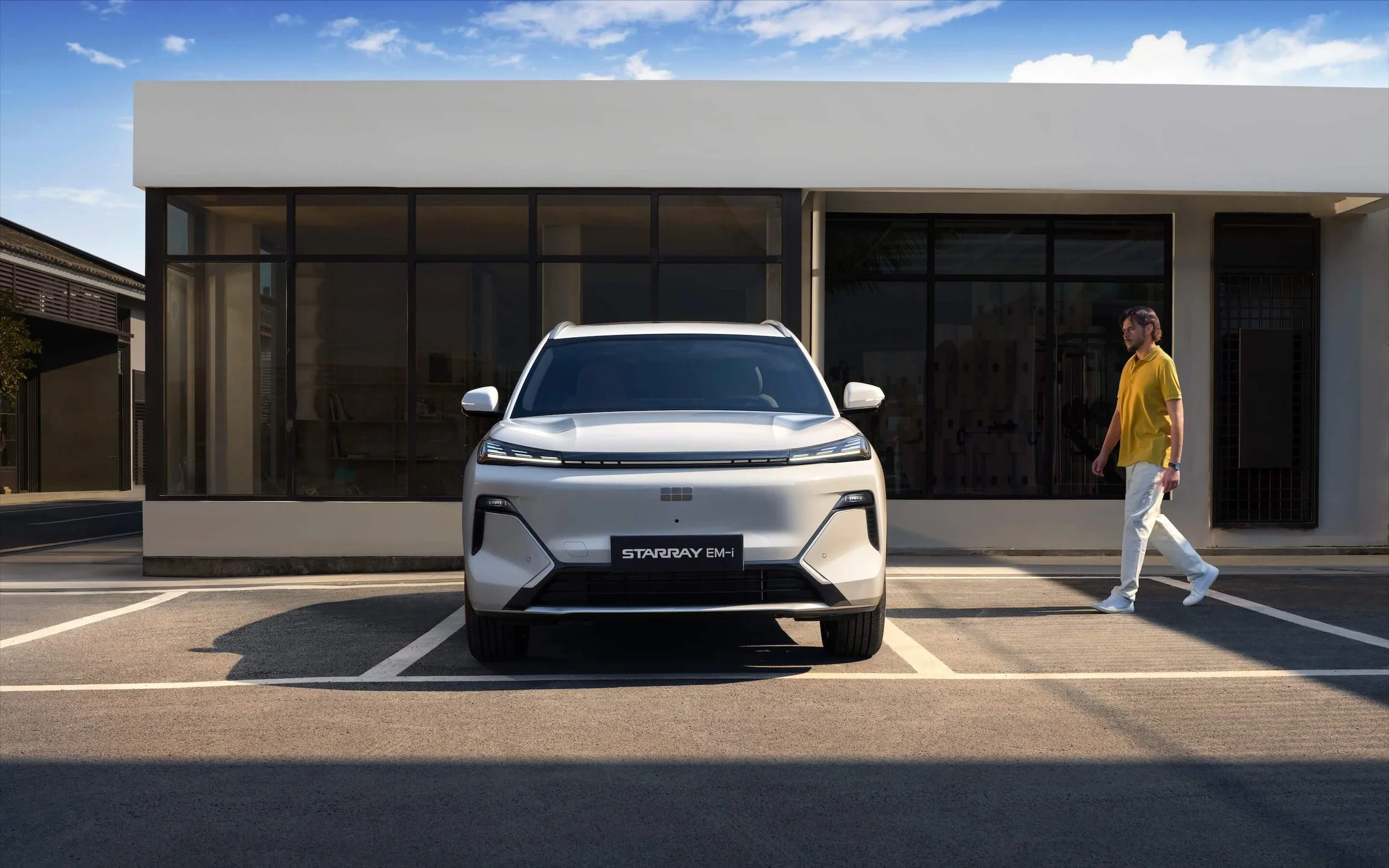 Coche eléctrico plateado estacionado en un espacio de aparcamiento frente a un edificio moderno, con un hombre caminando a su derecha.