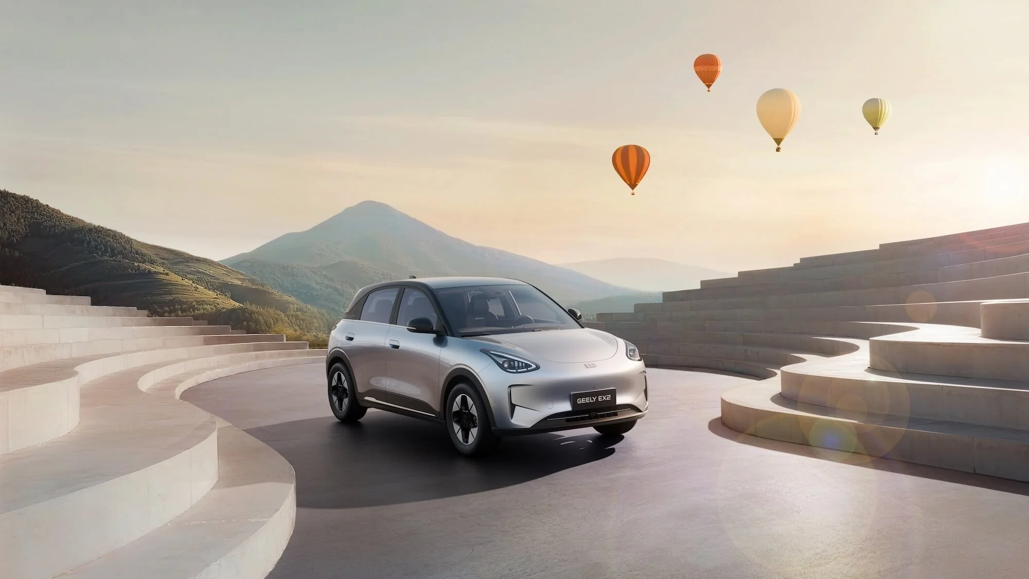 Coche eléctrico plateado en un espacio abierto con escaleras de cemento y montañas de fondo, con globos aerostáticos en el cielo al atardecer.