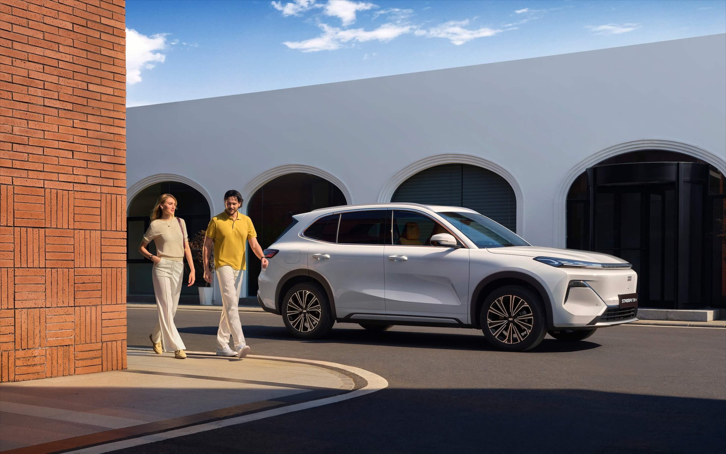 Un auto blanco estacionado en la calle frente a un edificio con ventanas arqueadas, con un hombre y una mujer caminando cerca del vehículo bajo un cielo despejado.