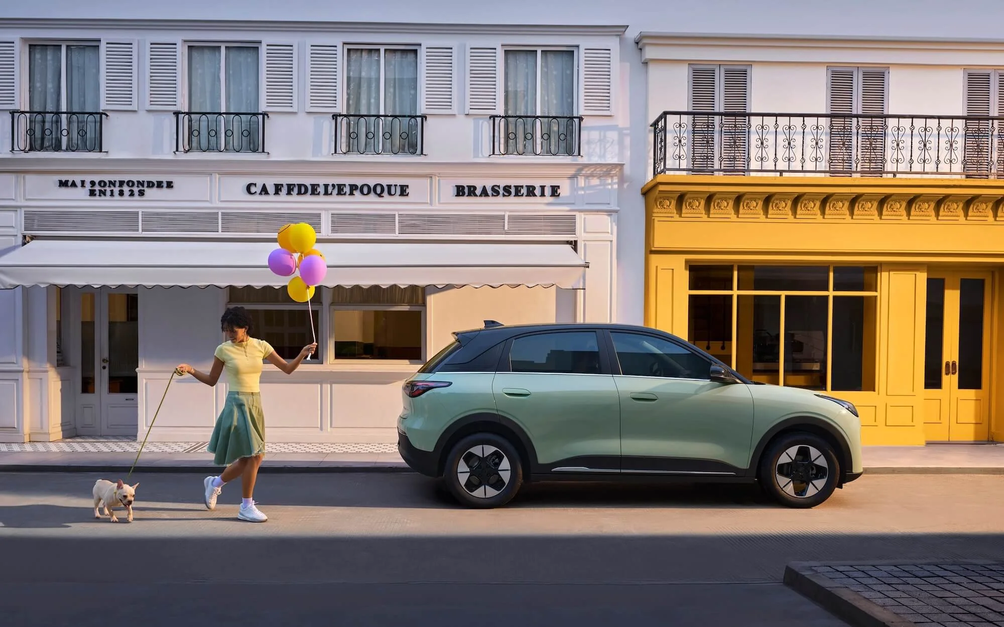 Mujer con vestido verde, paseando a su perro con globos en una calle con edificios blancos y amarillos.