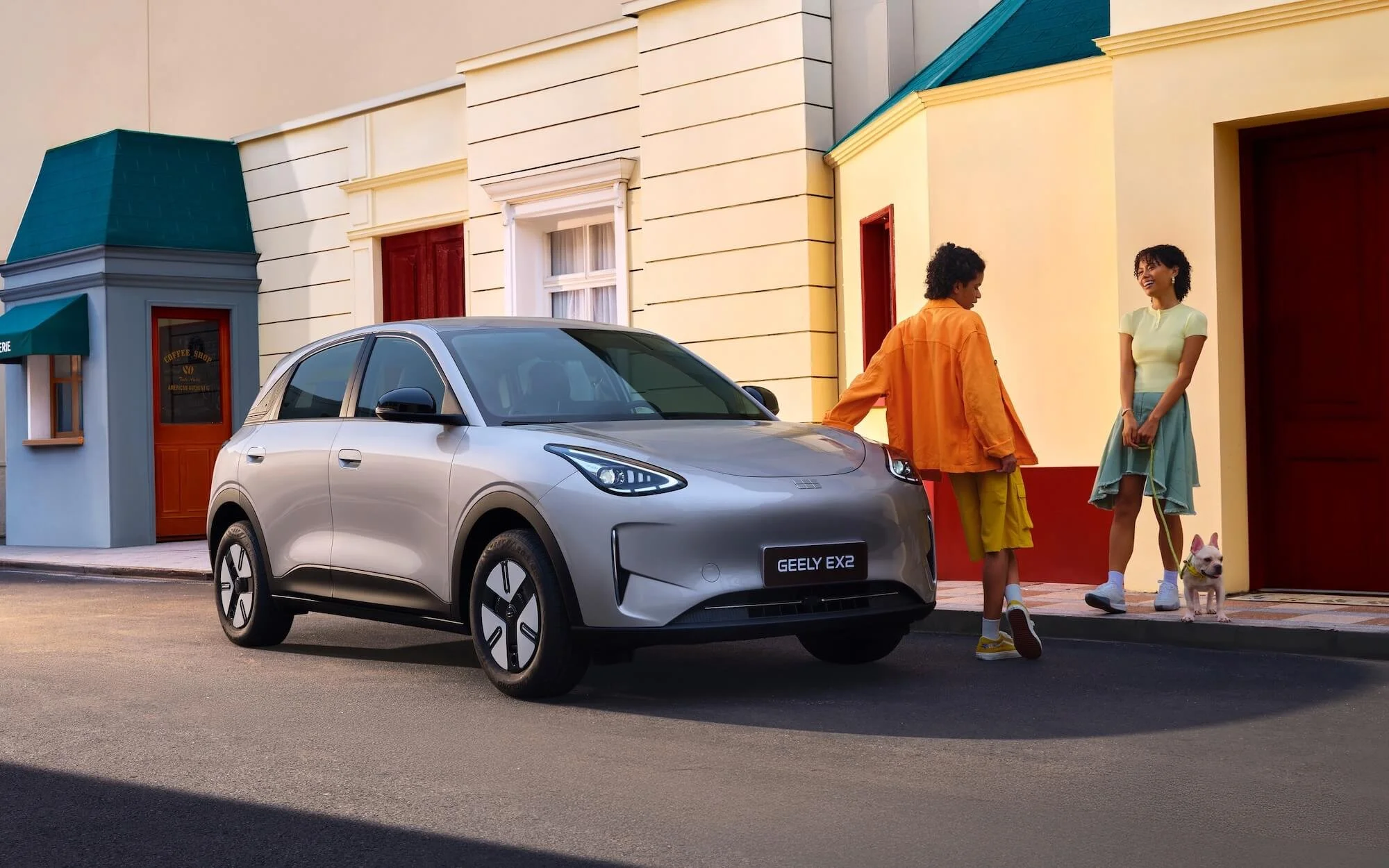 Auto eléctrico GEELY EX2 estacionado en una calle frente a una casa de colores vivos, dos mujeres conversando y un perro pequeño sentado en la acera.