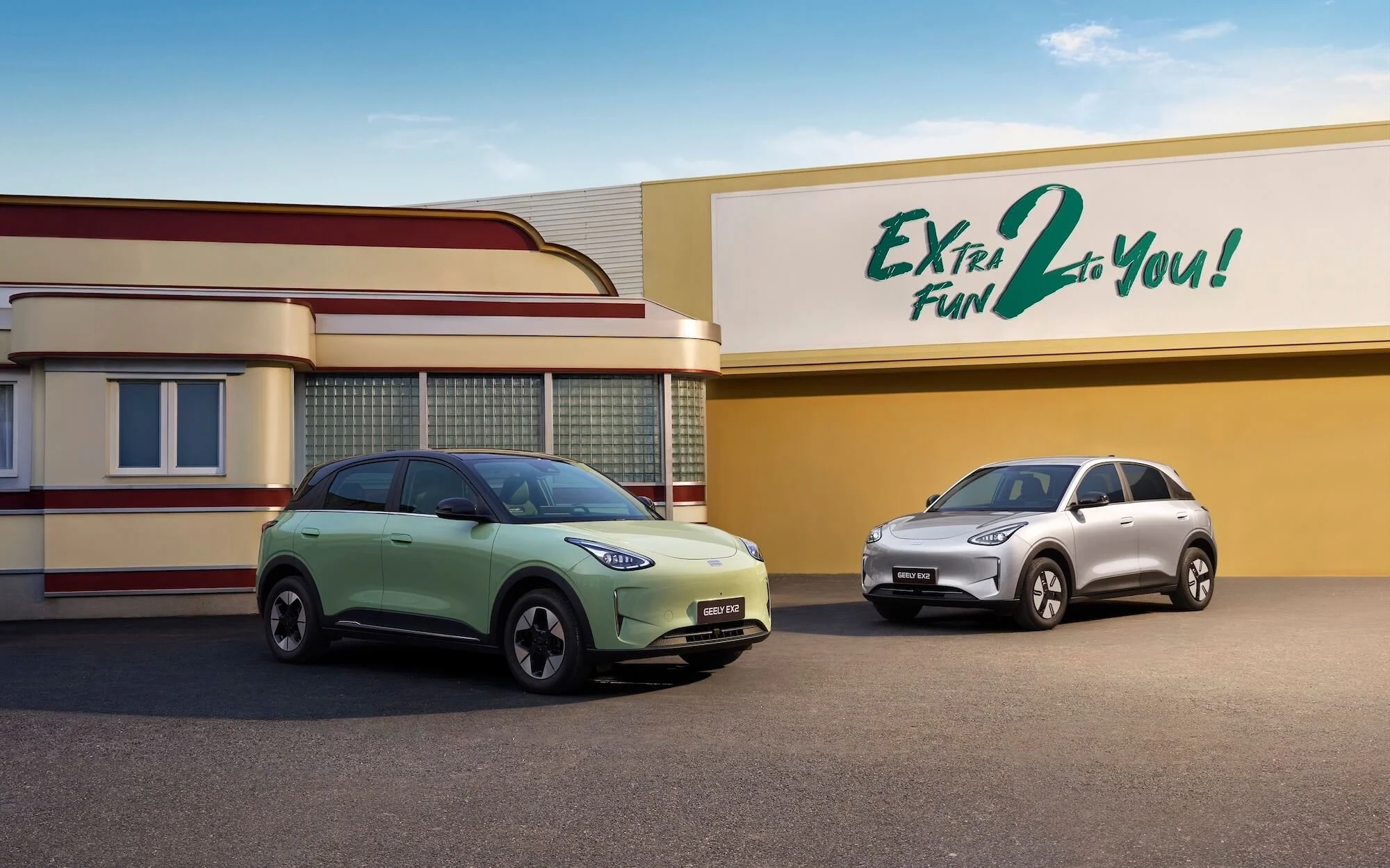 Dos autos eléctricos, uno verde y otro plateado, estacionados frente a un edificio con un cartel que dice 'Extra Fun 2 to You!'